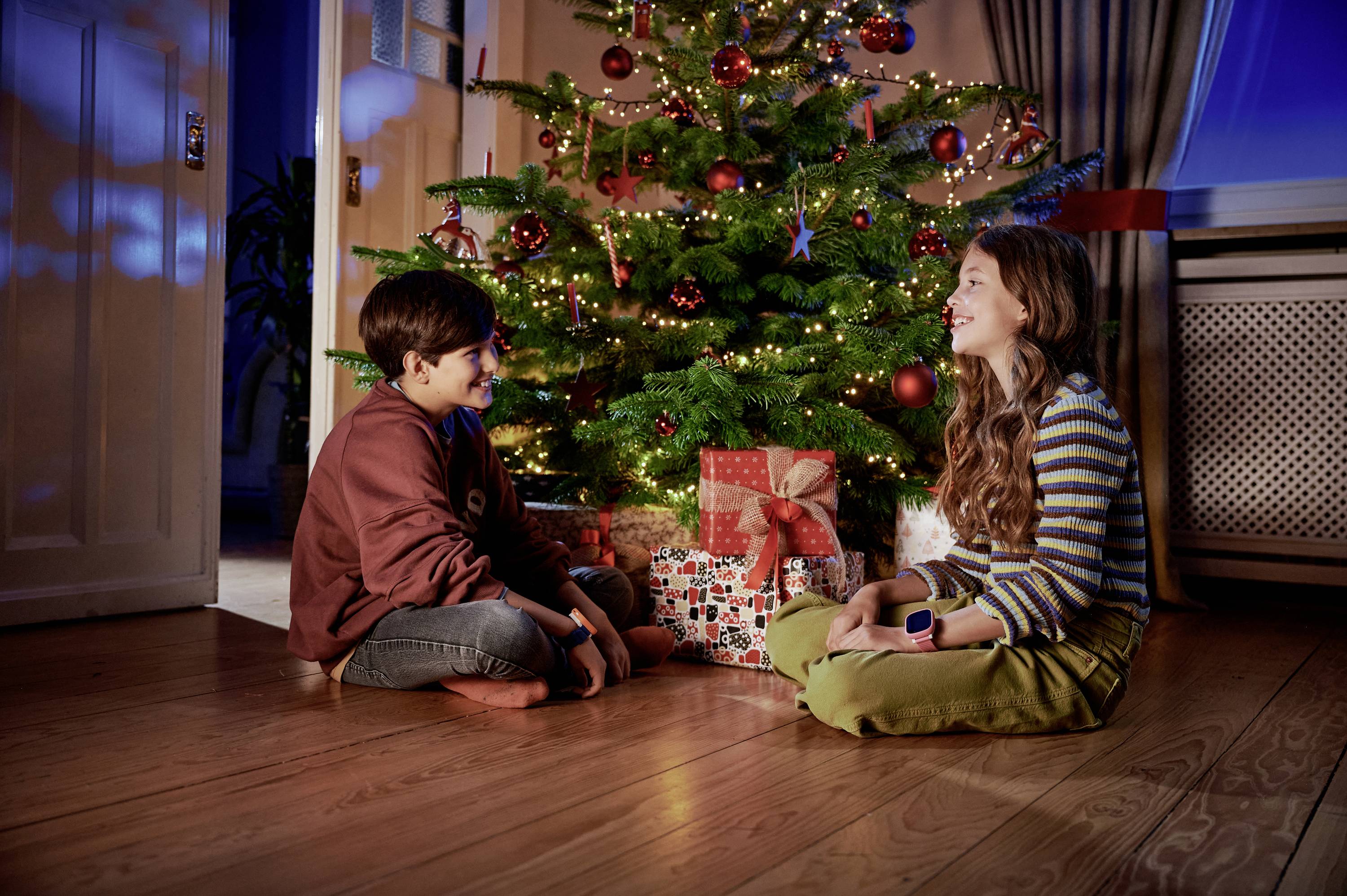 A boy and a girl are sitting smiling in front of a decorated Christmas tree with presents underneath. A festive atmosphere fills the room.