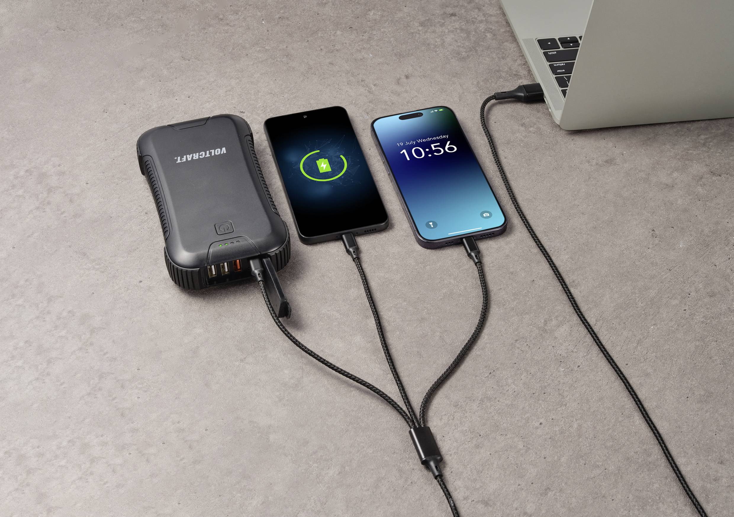 Four devices are connected on a concrete surface: a power bank, two charging smartphones, and a laptop.