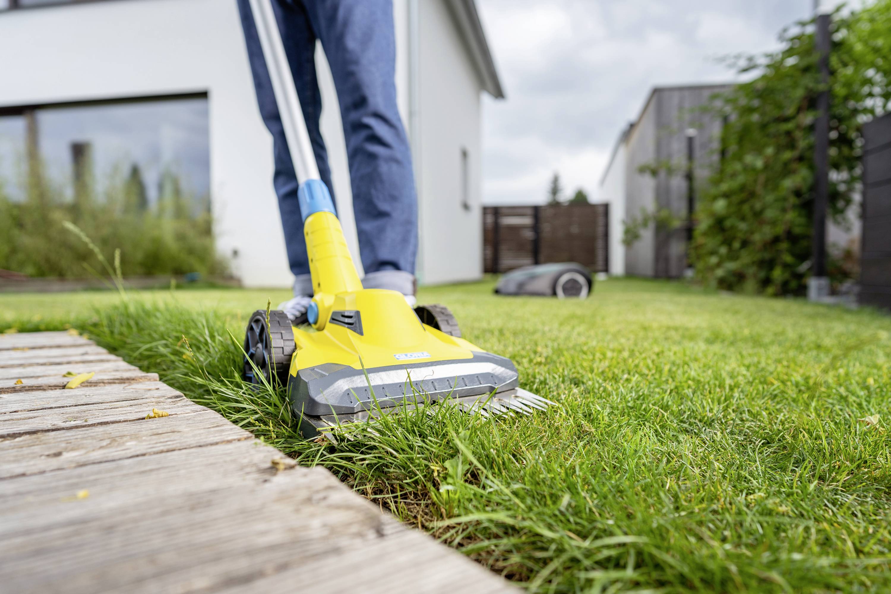 A person is using a yellow electric lawn trimmer to cut the lawn edges alongside a wooden path in a modern garden.