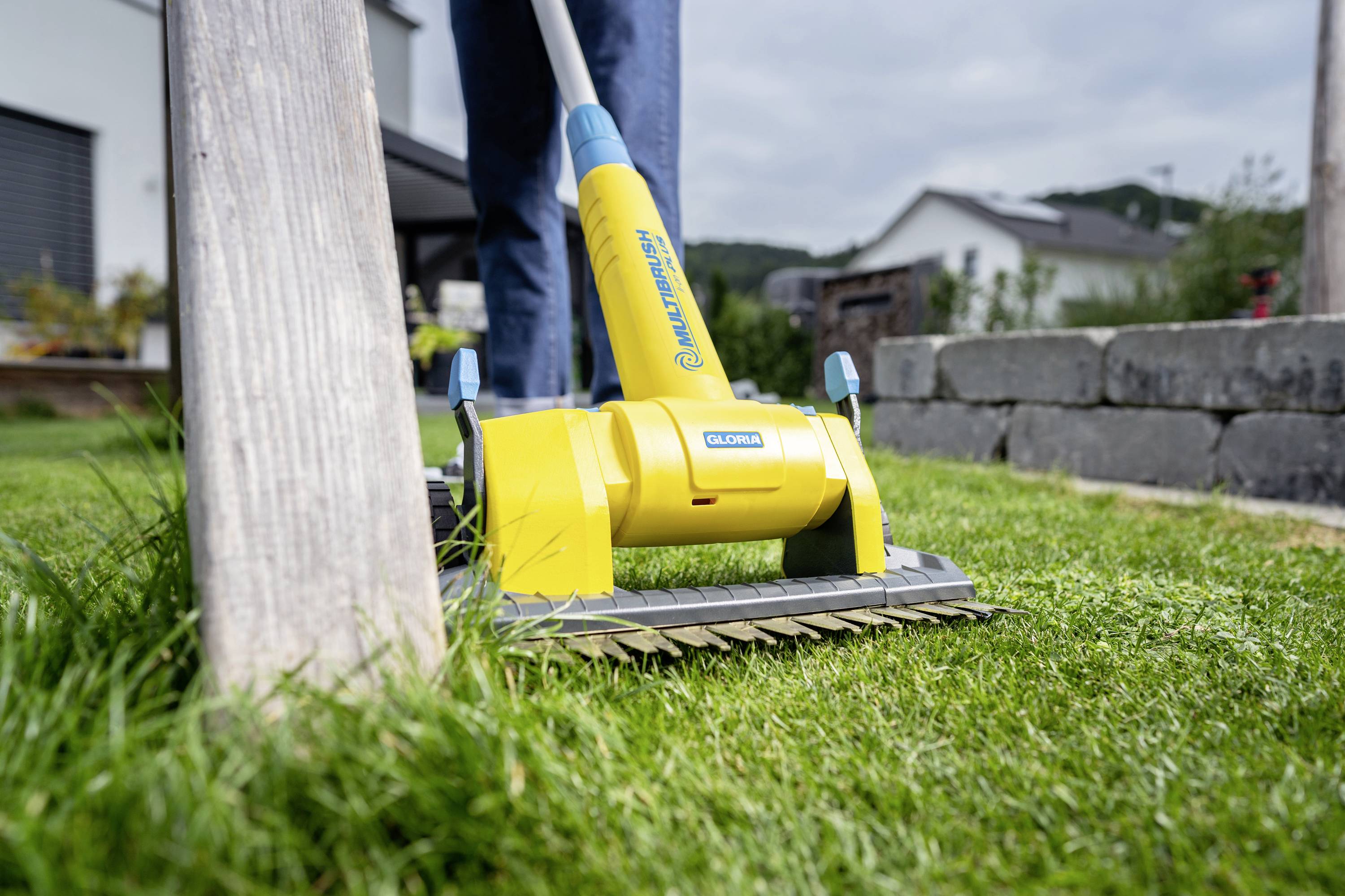 Close-up of a yellow electric lawnmower head moving across a green grassy area. Houses are visible in the background.
