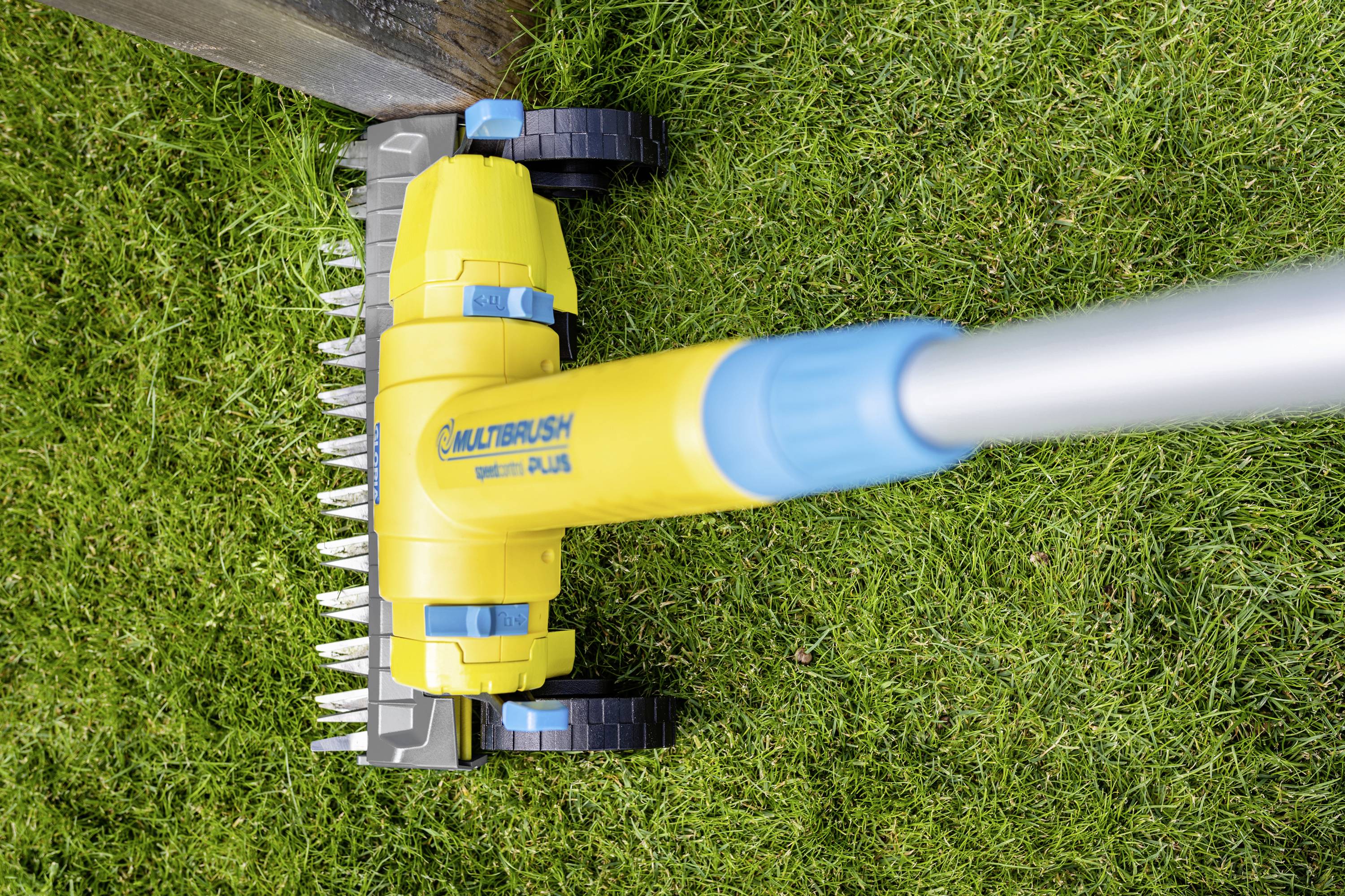 A handheld device with a yellow brush and wheels trims grass along an edge on a green lawn.