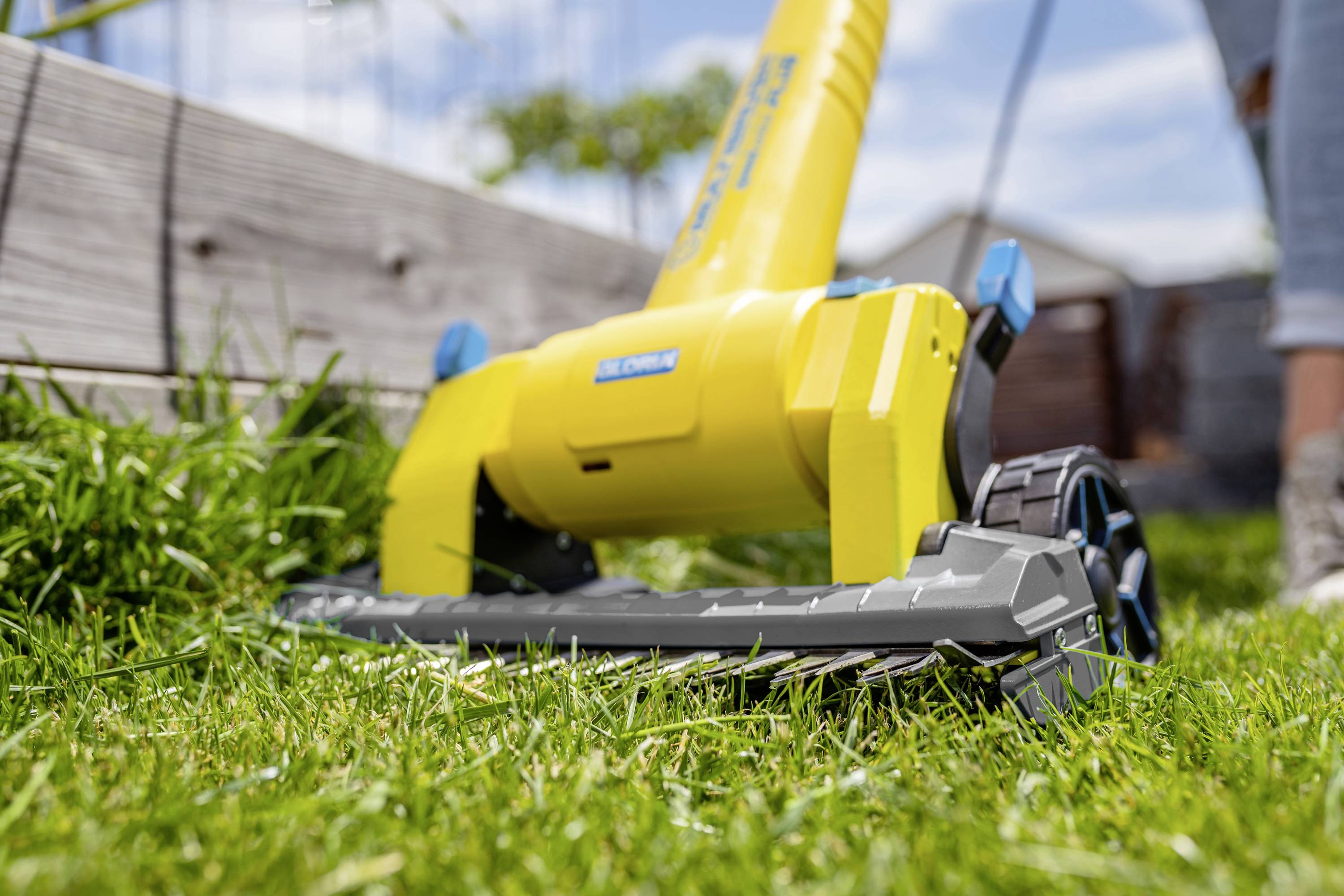 The foreground shows a yellow garden tool on green grass, most likely a lawnmower. In the background are wooden boards and blurred plants.