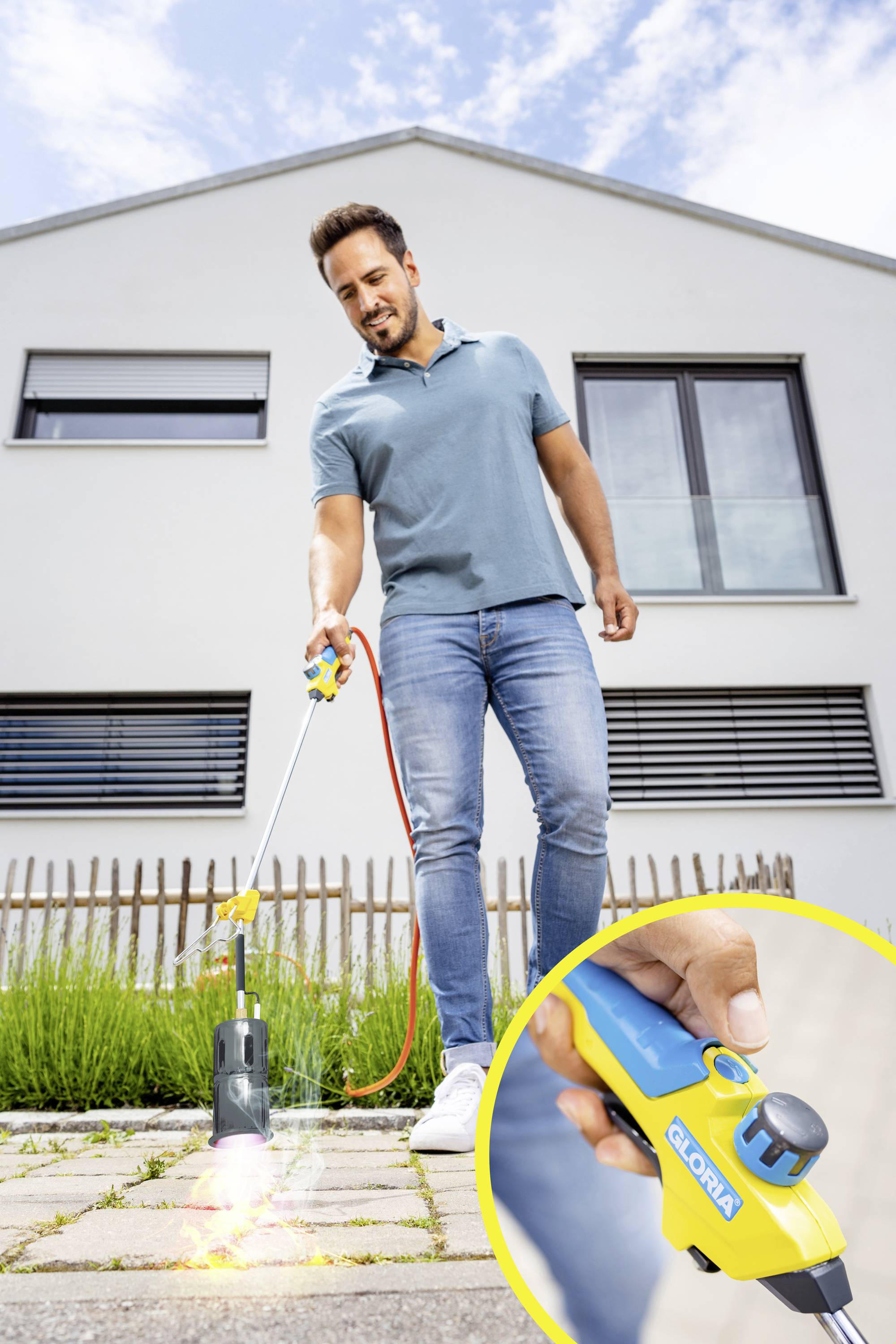 A man is using a weed burning device on a pavement in front of a modern house. Close-up of the device handle in the bottom right.