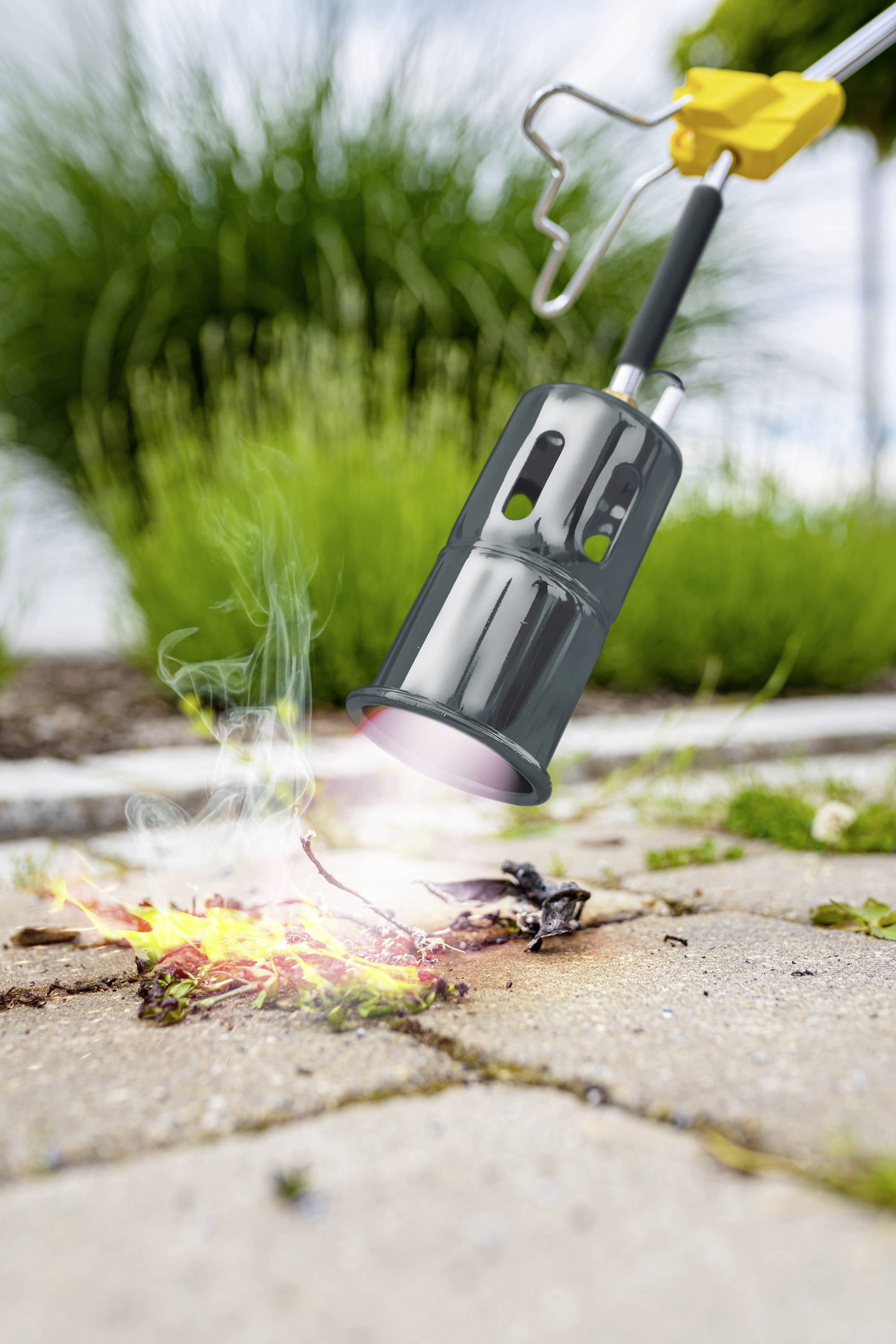 A weed burner destroys plants on a paved path. Smoke rises, the surroundings are green and summery.