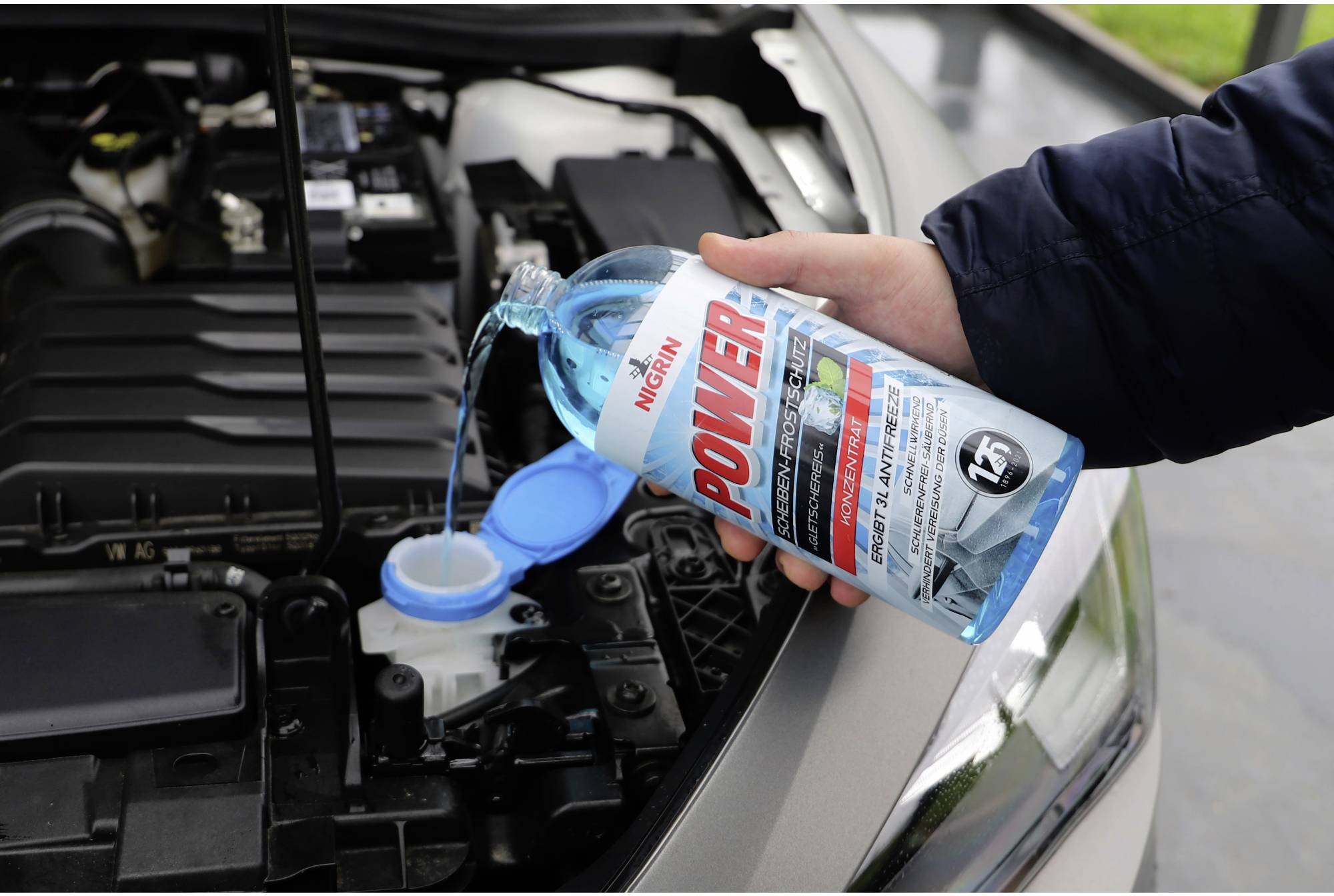 A person is filling windscreen washer fluid into the reservoir of a car. An open bonnet reveals parts of the engine.