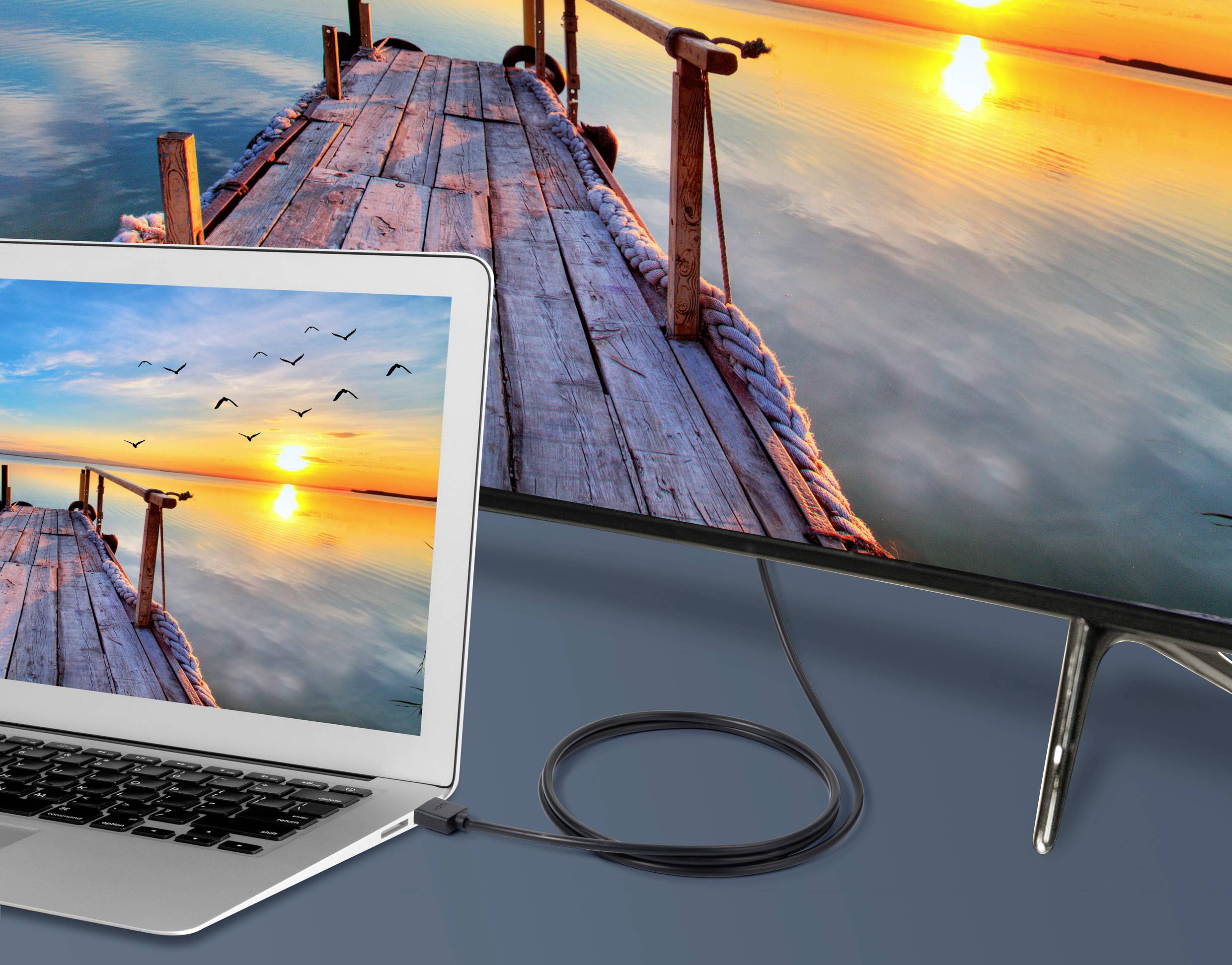 A laptop displays a jetty leading into a sunset over a lake. The laptop is connected to a larger screen showing the same image.