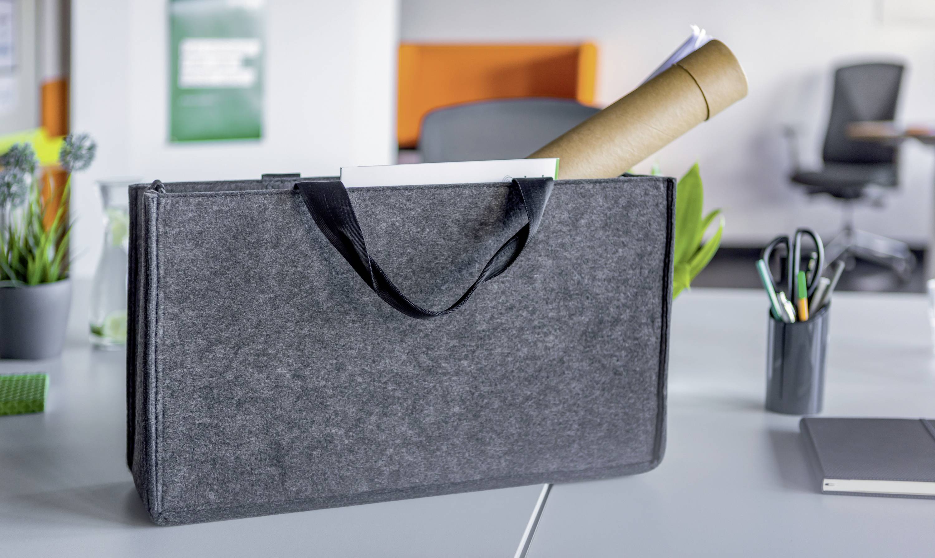 A grey document folder lies open on a desk. Paper rolls and documents are inside it. Office supplies and a blurred chair are visible in the background.