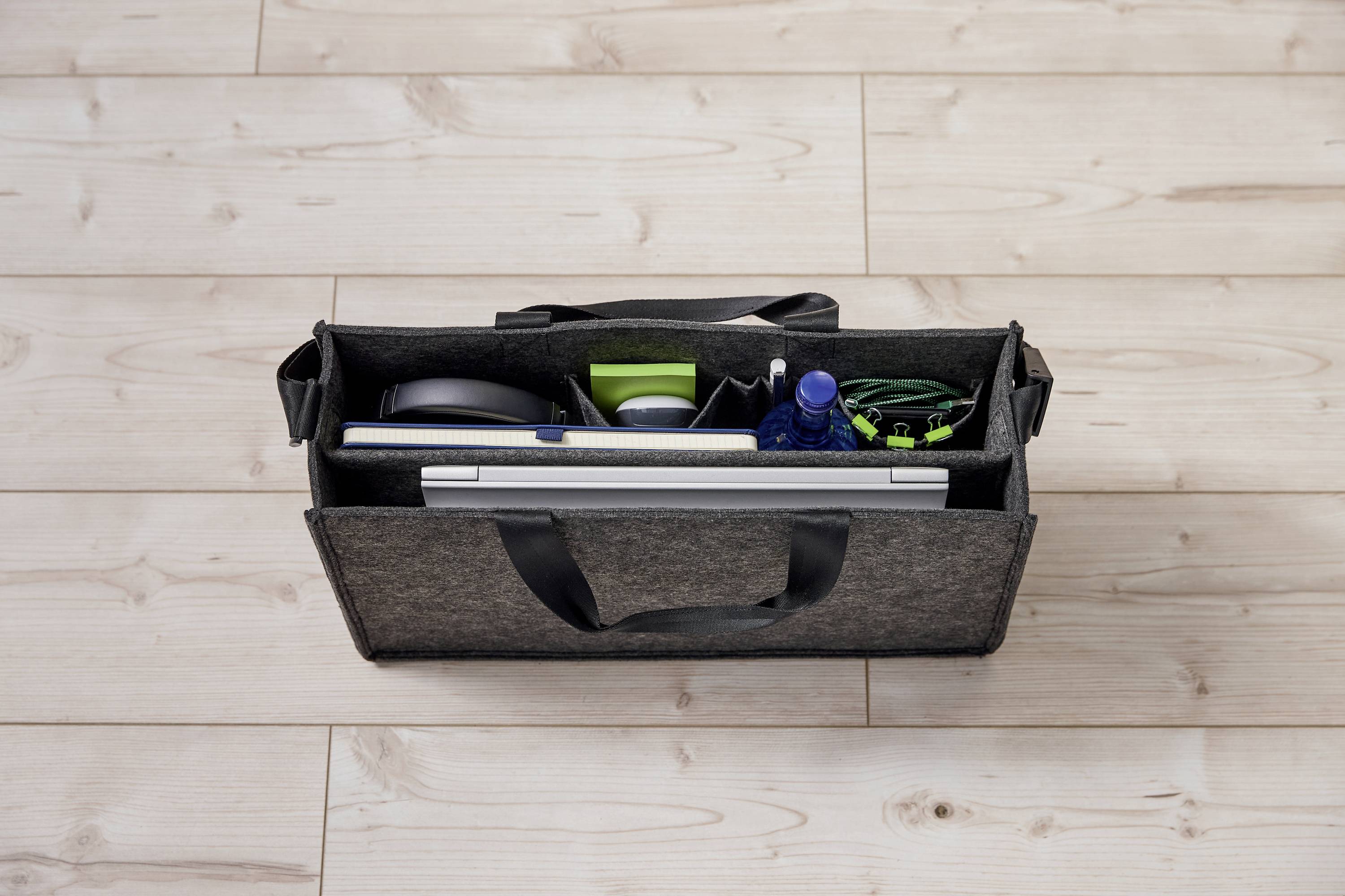 A grey bag on a wooden floor, opened to reveal a laptop, headphones, water bottle, and cables, displaying travel or work essentials.