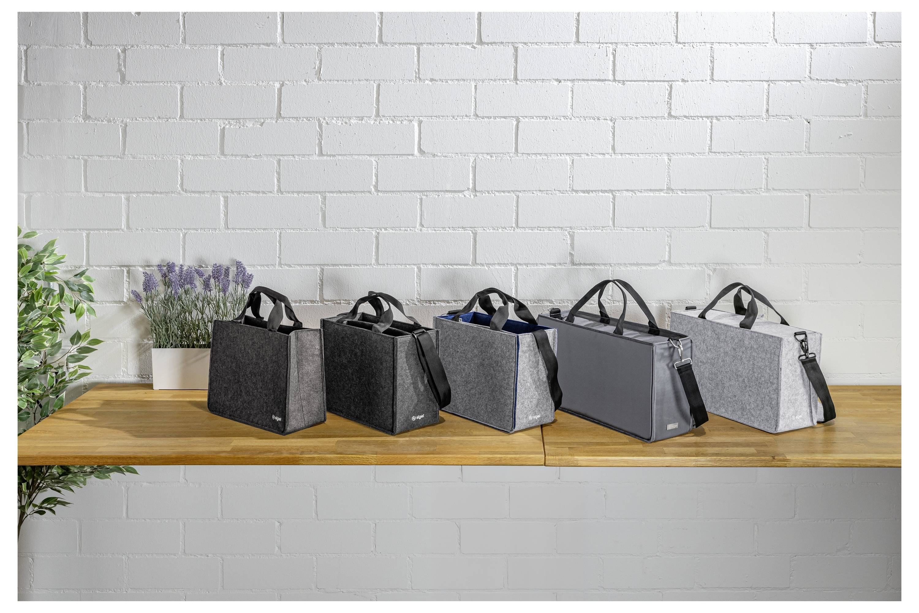 Five insulated tote bags in black, gray, and light gray lined up on a wooden shelf against a white brick wall.
