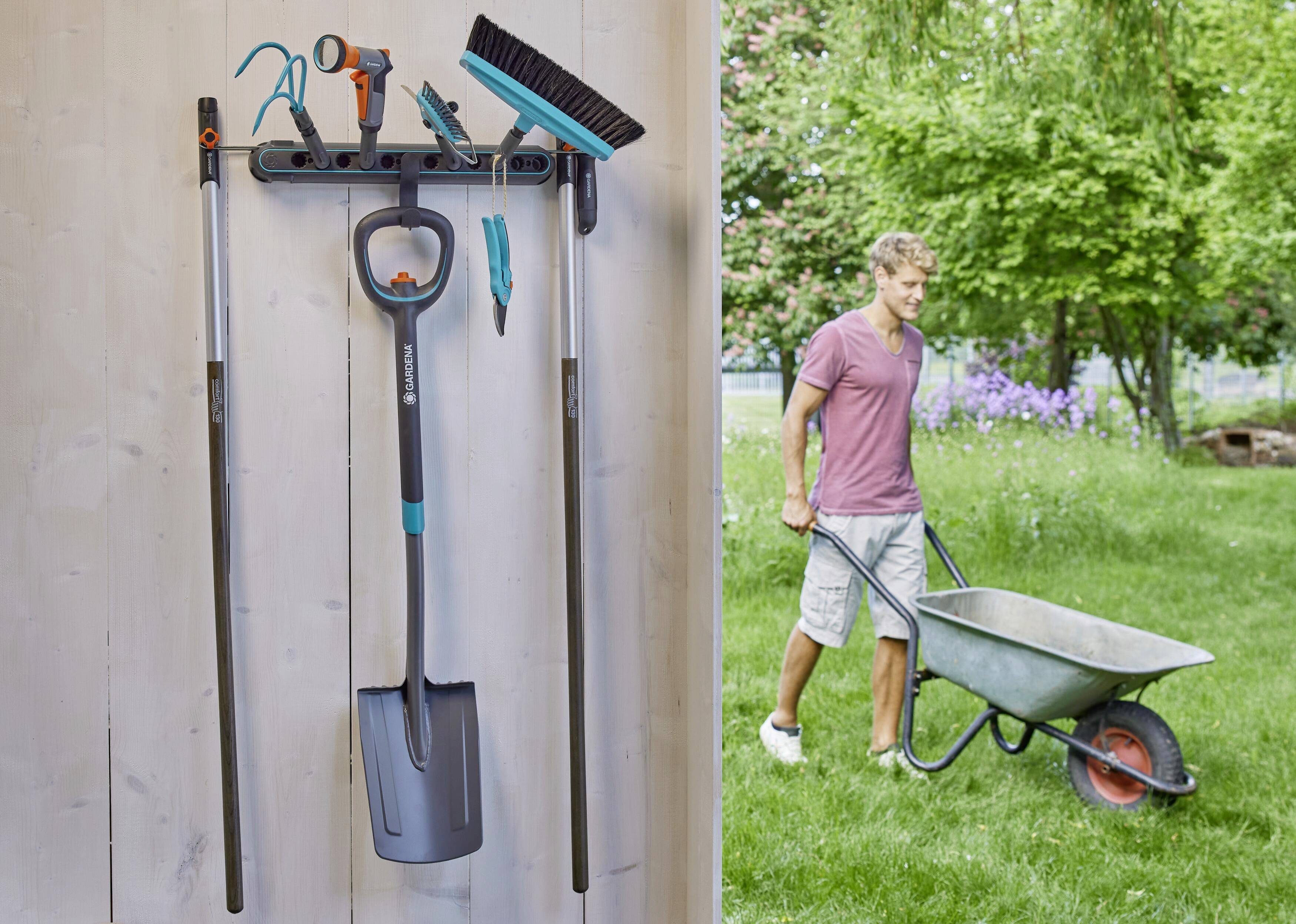 'A wall-mounted rack with garden tools on the left; a man in shorts and a t-shirt pushing a wheelbarrow across a green lawn on the right.'