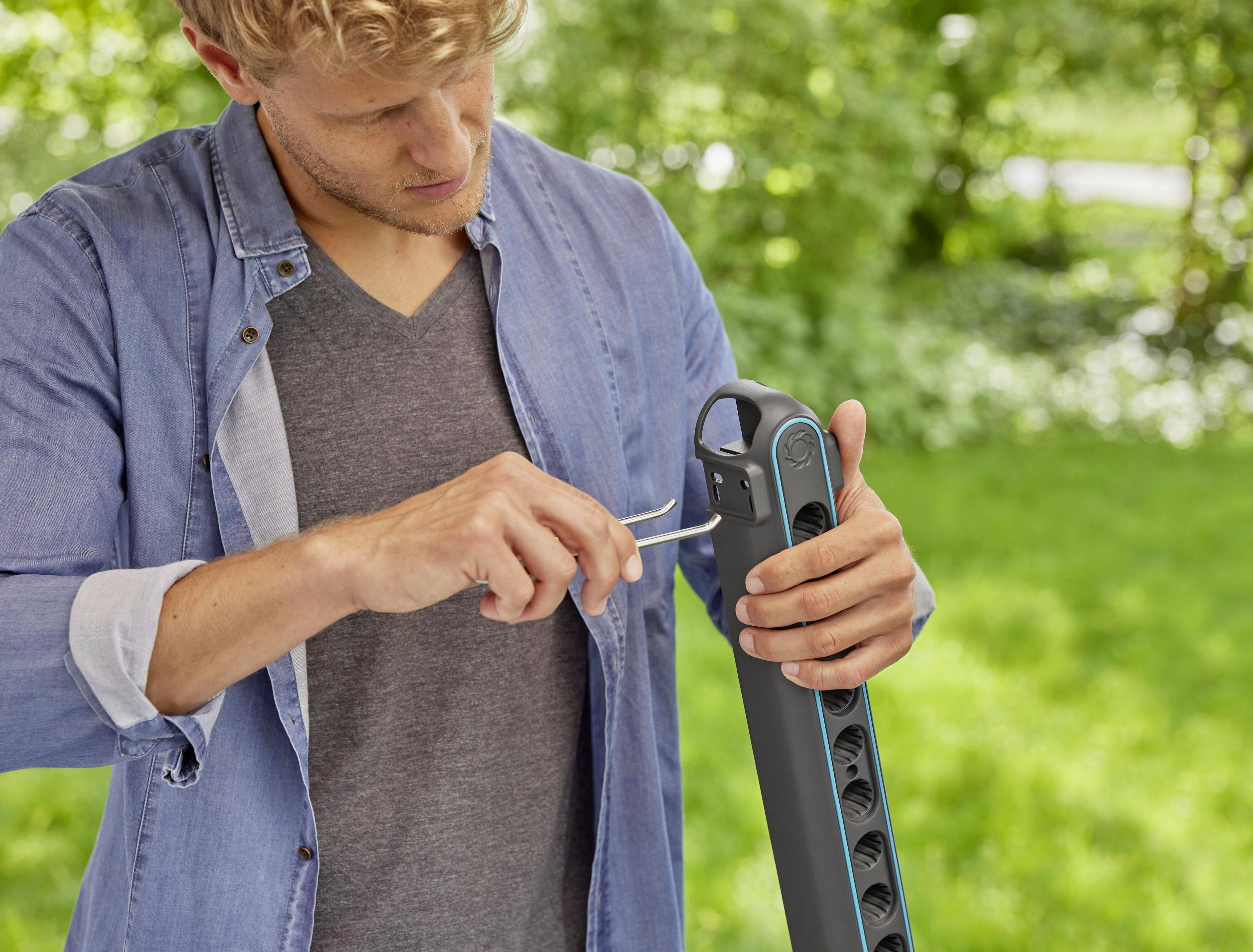 A man is using a screwdriver in the garden to repair a black, vertical garden device. Green foliage is visible in the background.