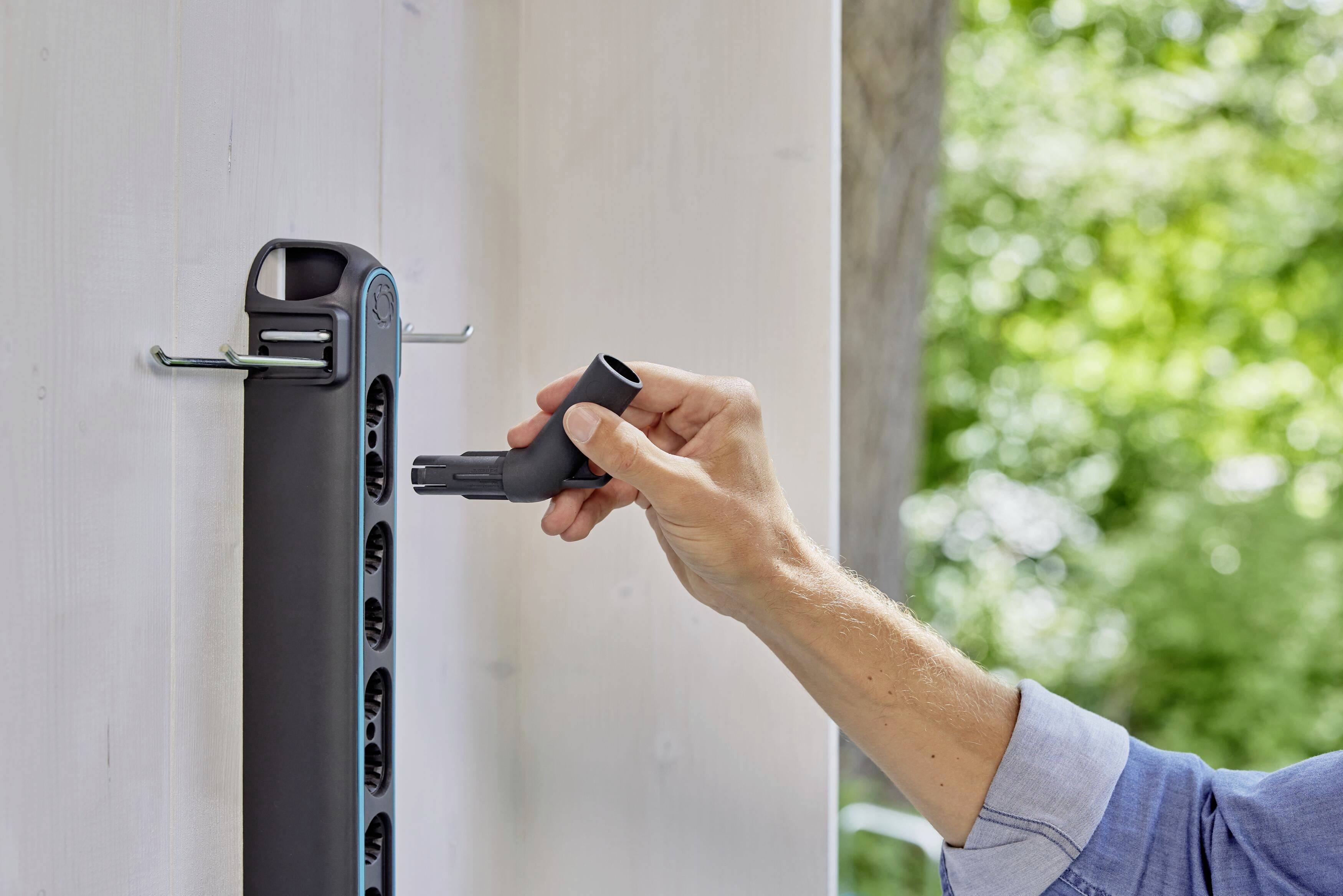A person is hanging a garden hose on a holder attached to a white wall, with green vegetation visible in the background.