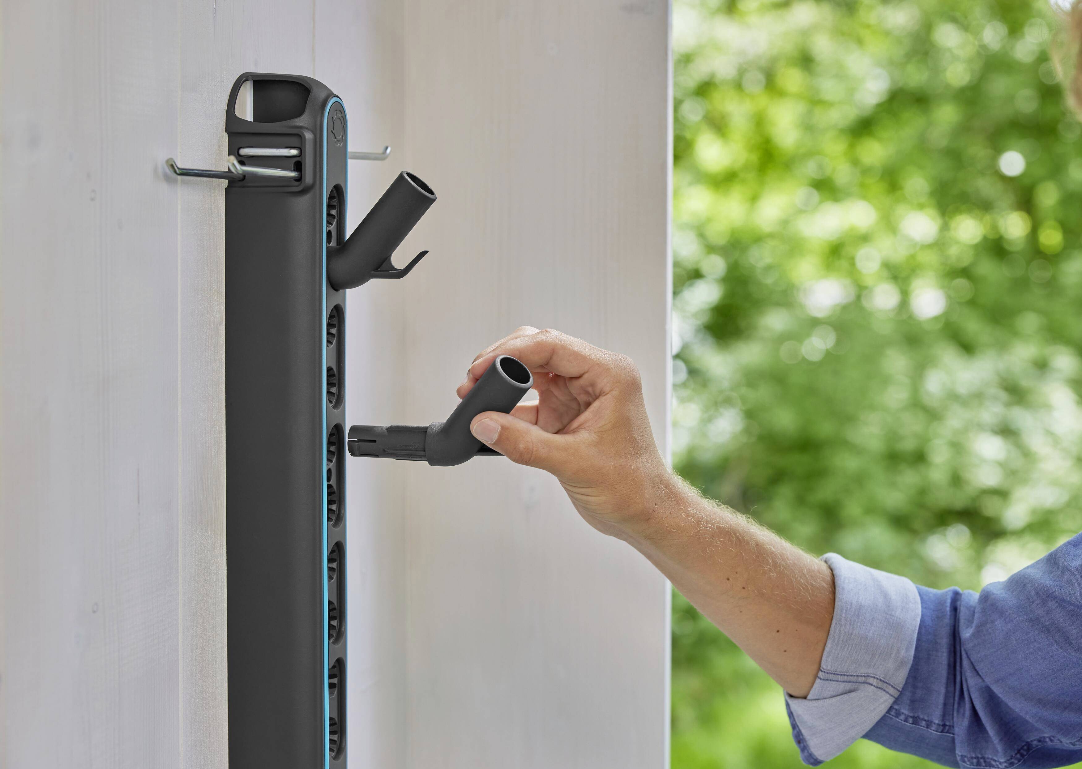 A person is attaching a tool to a storage rail with multiple holders, with green vegetation visible in the background.