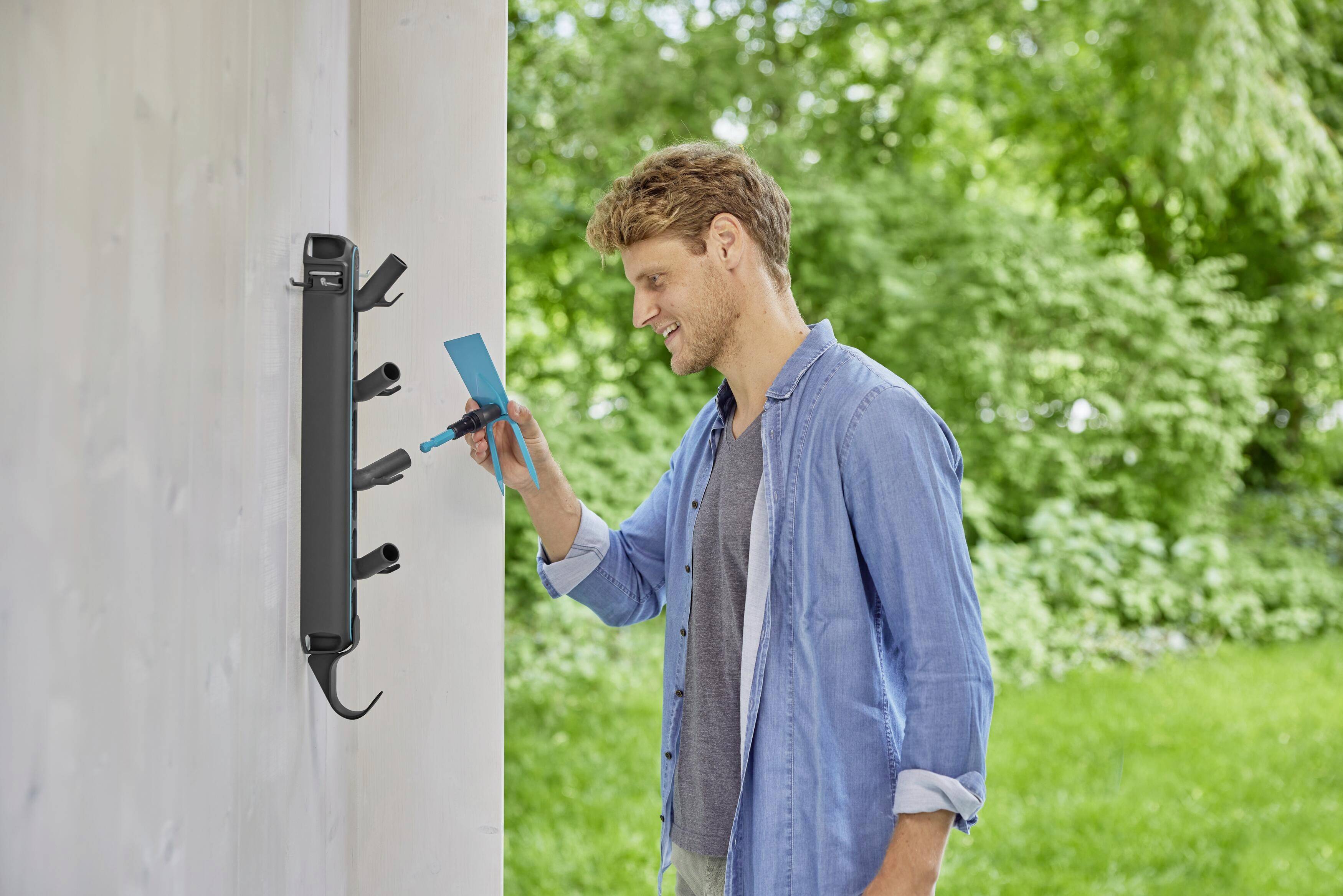 A man plugs a smartphone into an outdoor charging station on a wall in a green outdoor setting. Smiling, wearing a light blue shirt, on a sunny day.