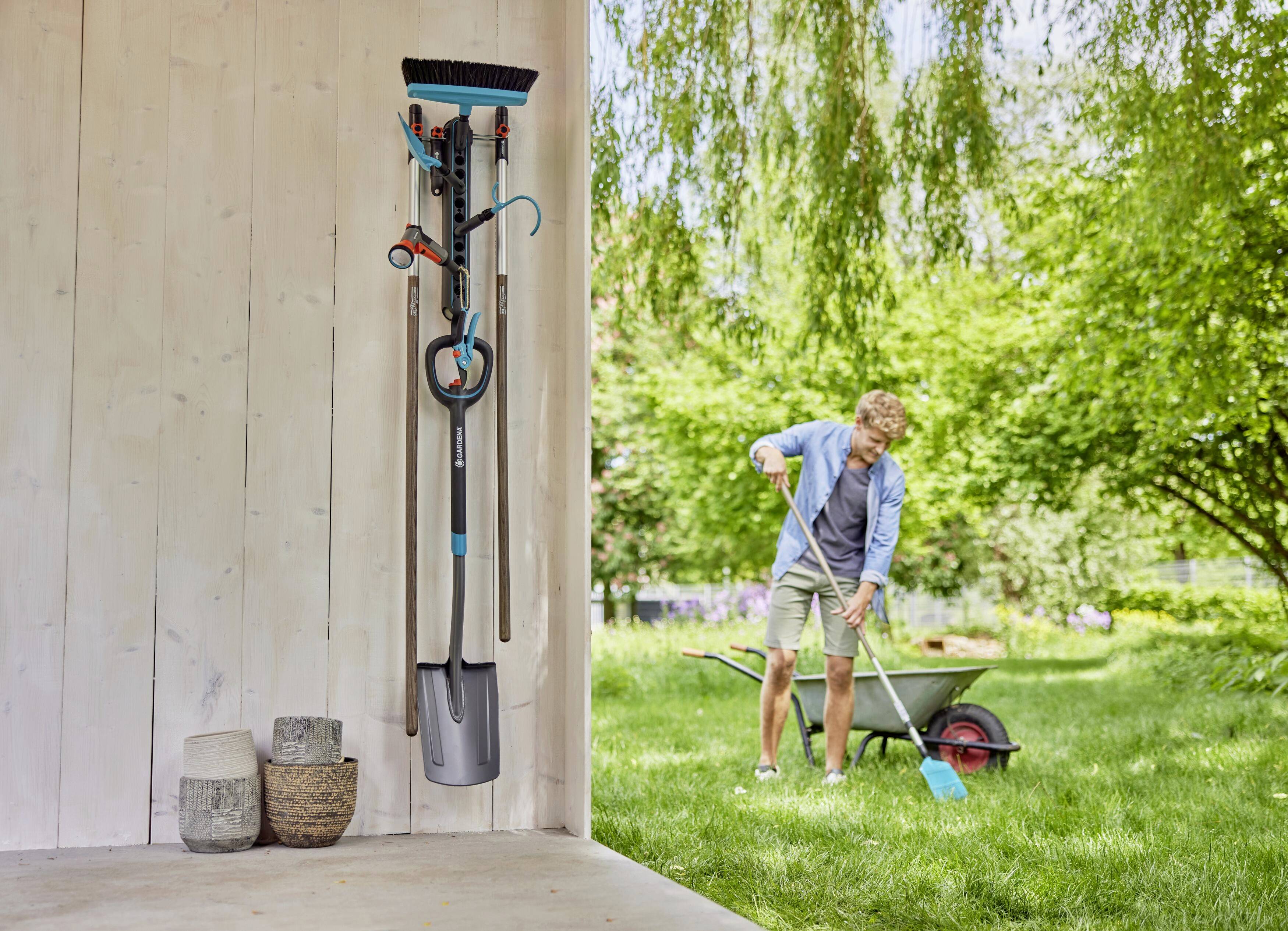 A man is working in the garden next to a light wooden wall with garden tools hanging on it. A wheelbarrow stands in the background.