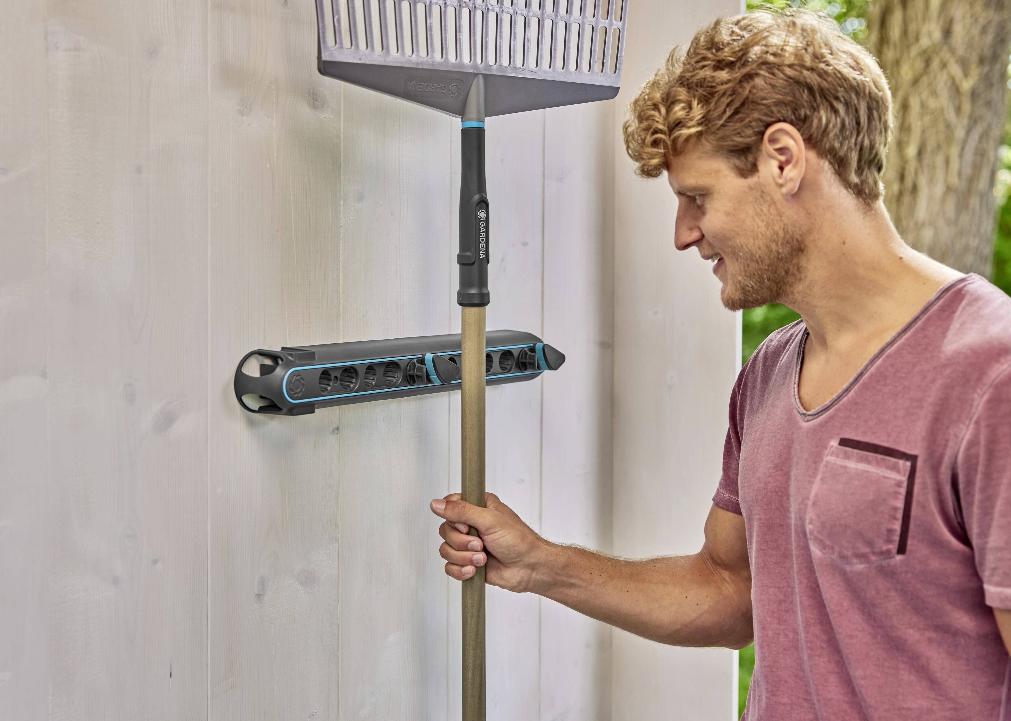 A man is hanging a rake on a wall bracket in a shed. The bracket is suitable for storing garden tools.