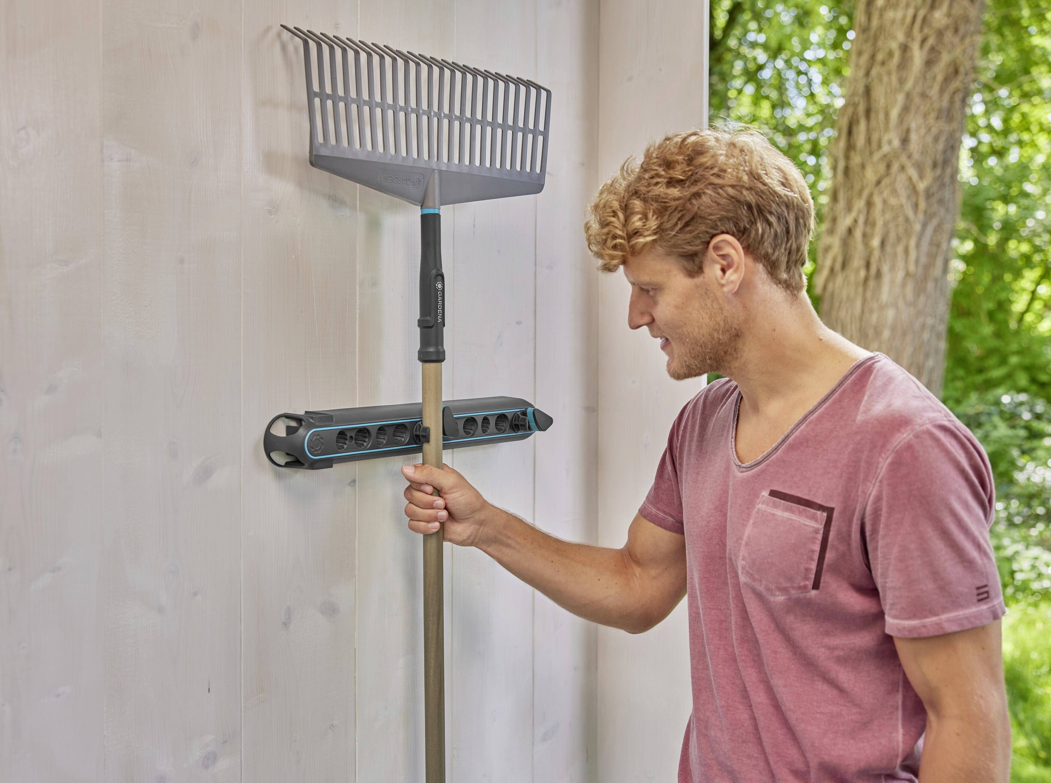 A man in a pink T-shirt is hanging a rake on a wall-mounted bracket attached to a white wall outdoors.