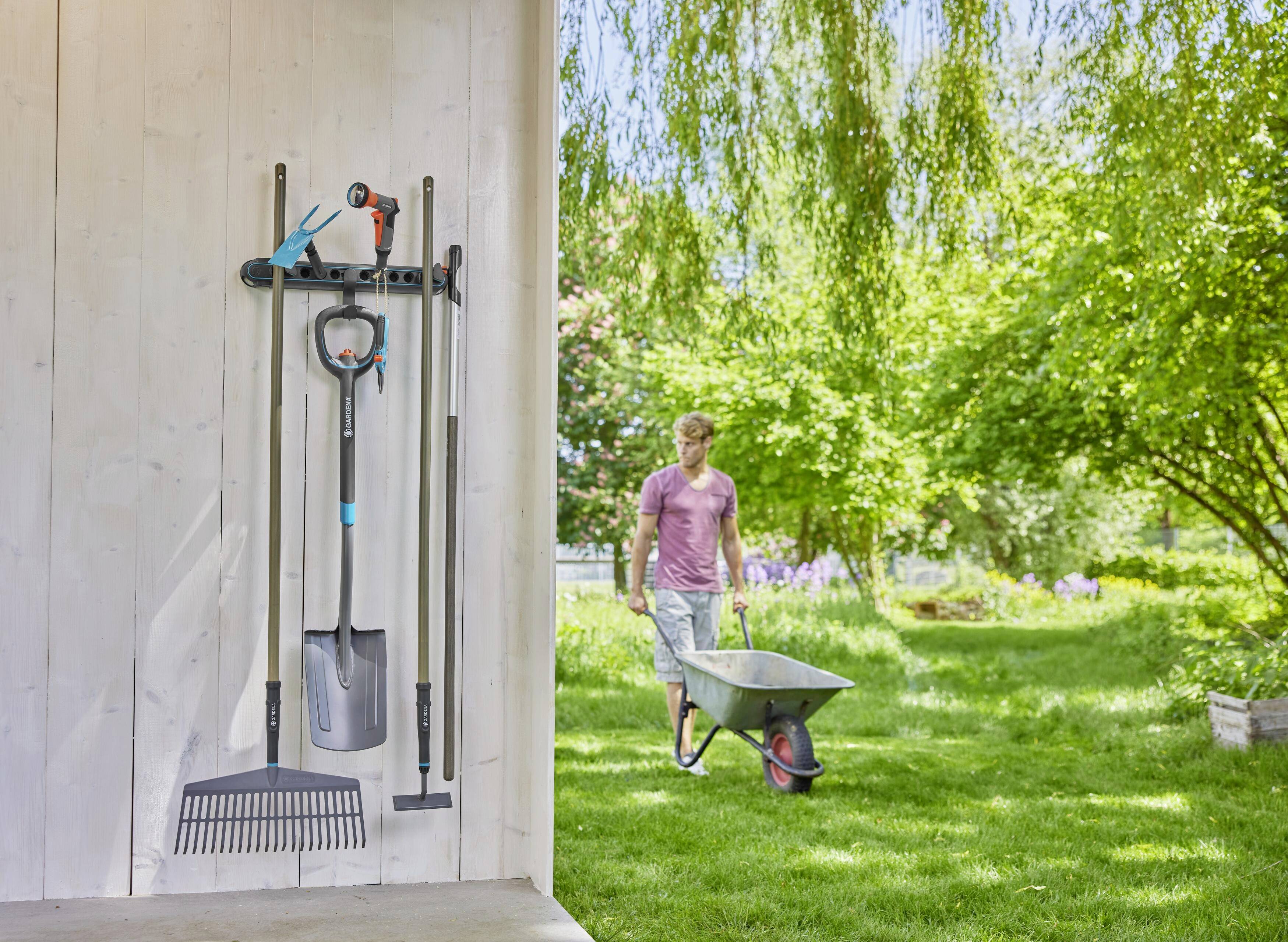 A man is pushing a wheelbarrow in the garden. Garden tools, including a rake and spade, are hanging on the wall. Sunshine and green trees are in the background.