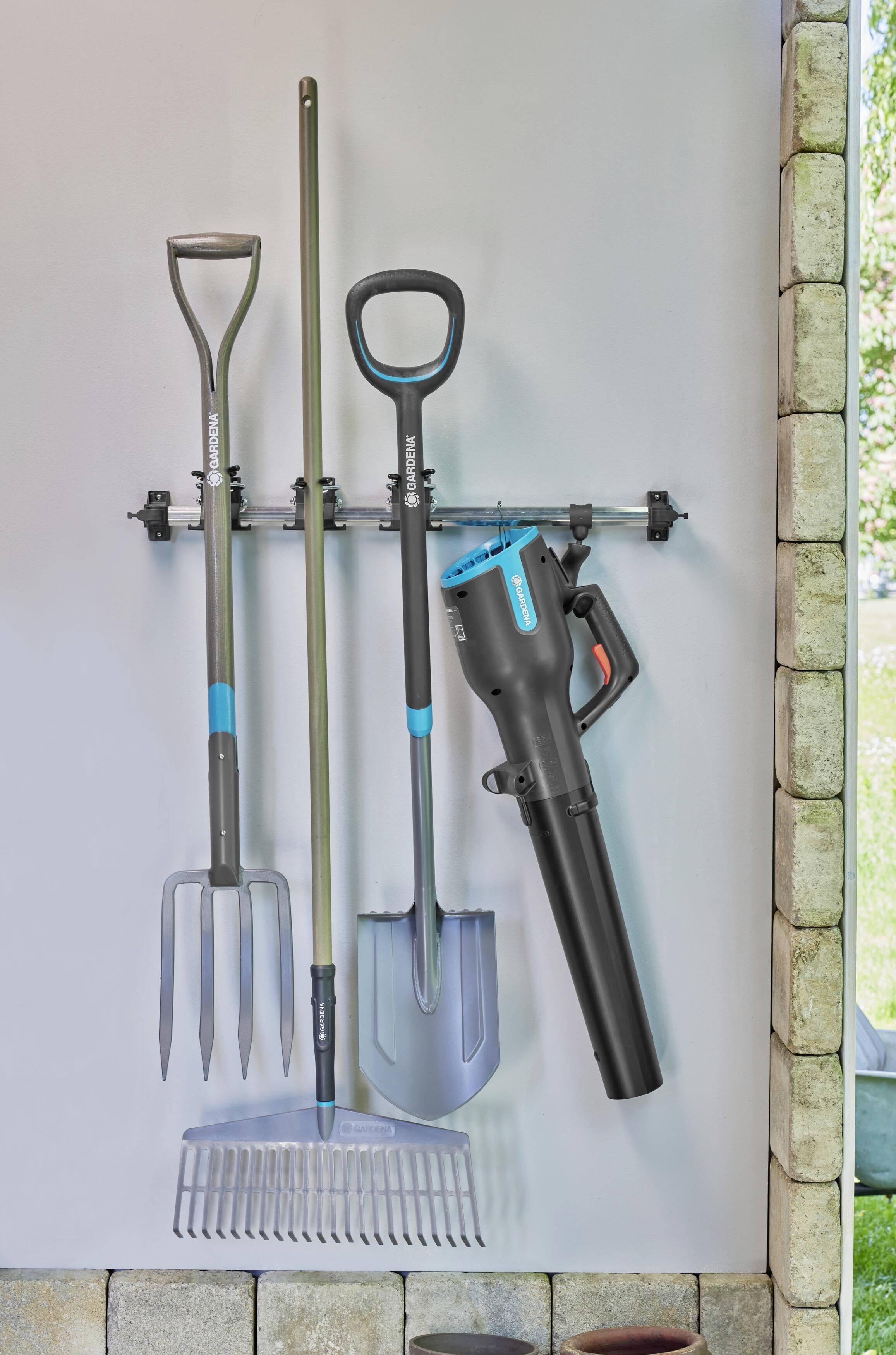 Garden tools hang on a wall mount. Visible are a pitchfork, a rake, a shovel, and a leaf blower.