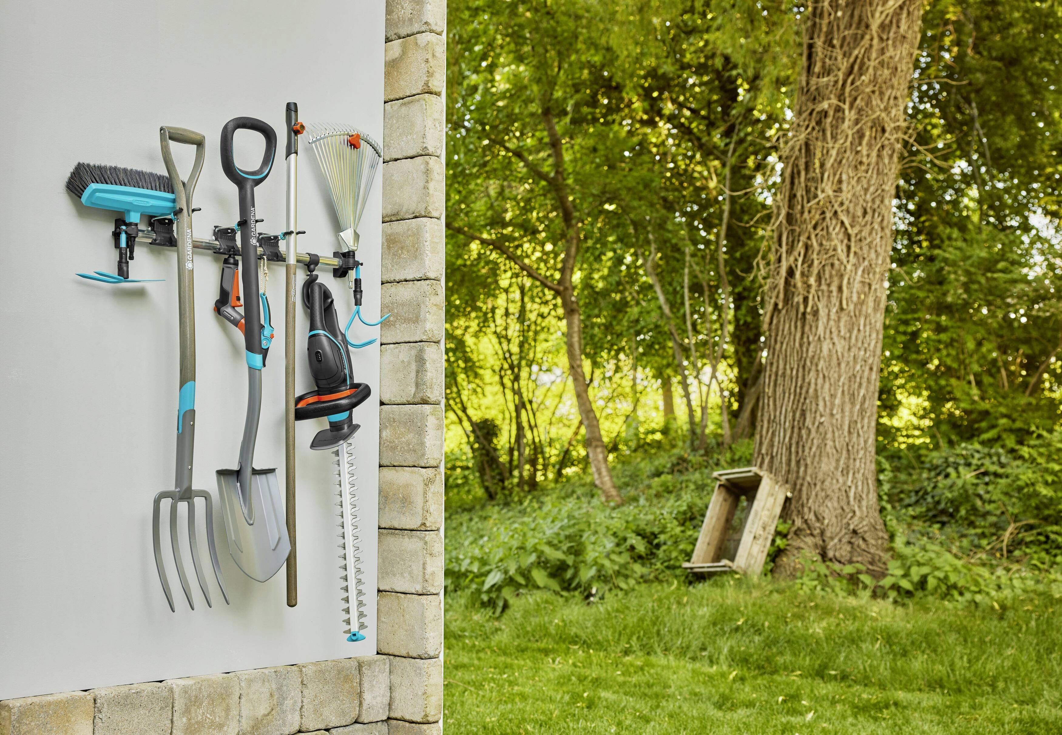 Garden tools such as brooms, rakes and hedge trimmers are neatly hung on a wall mounting. In the background, a tree and green lawn.