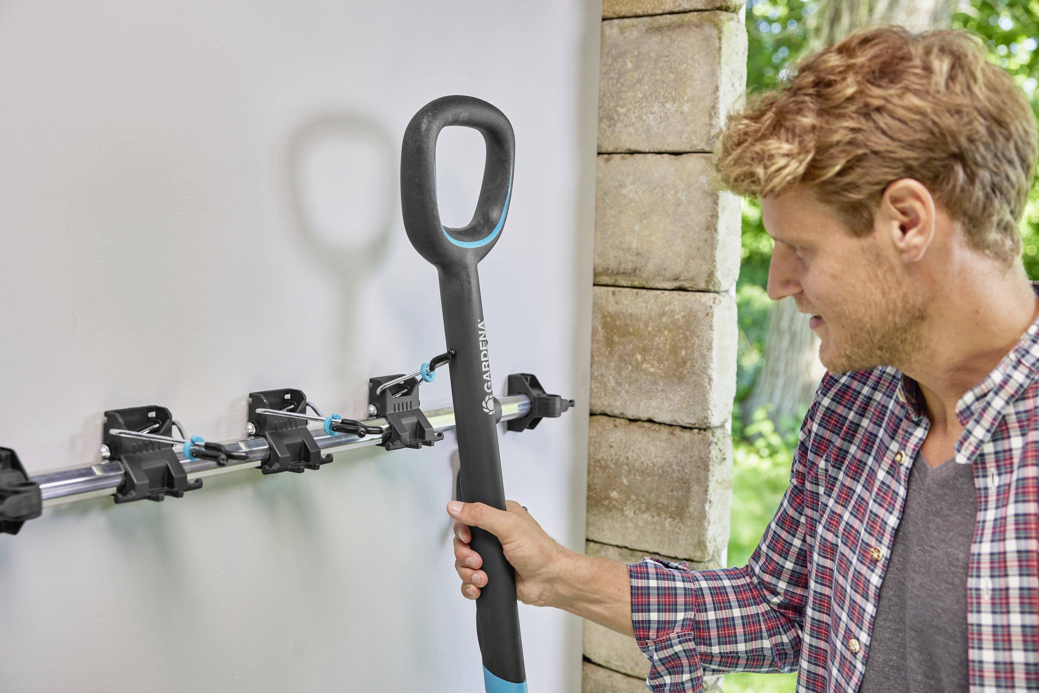 A man is hanging a shovel on a wall mounting system in a room with a view of trees outside.