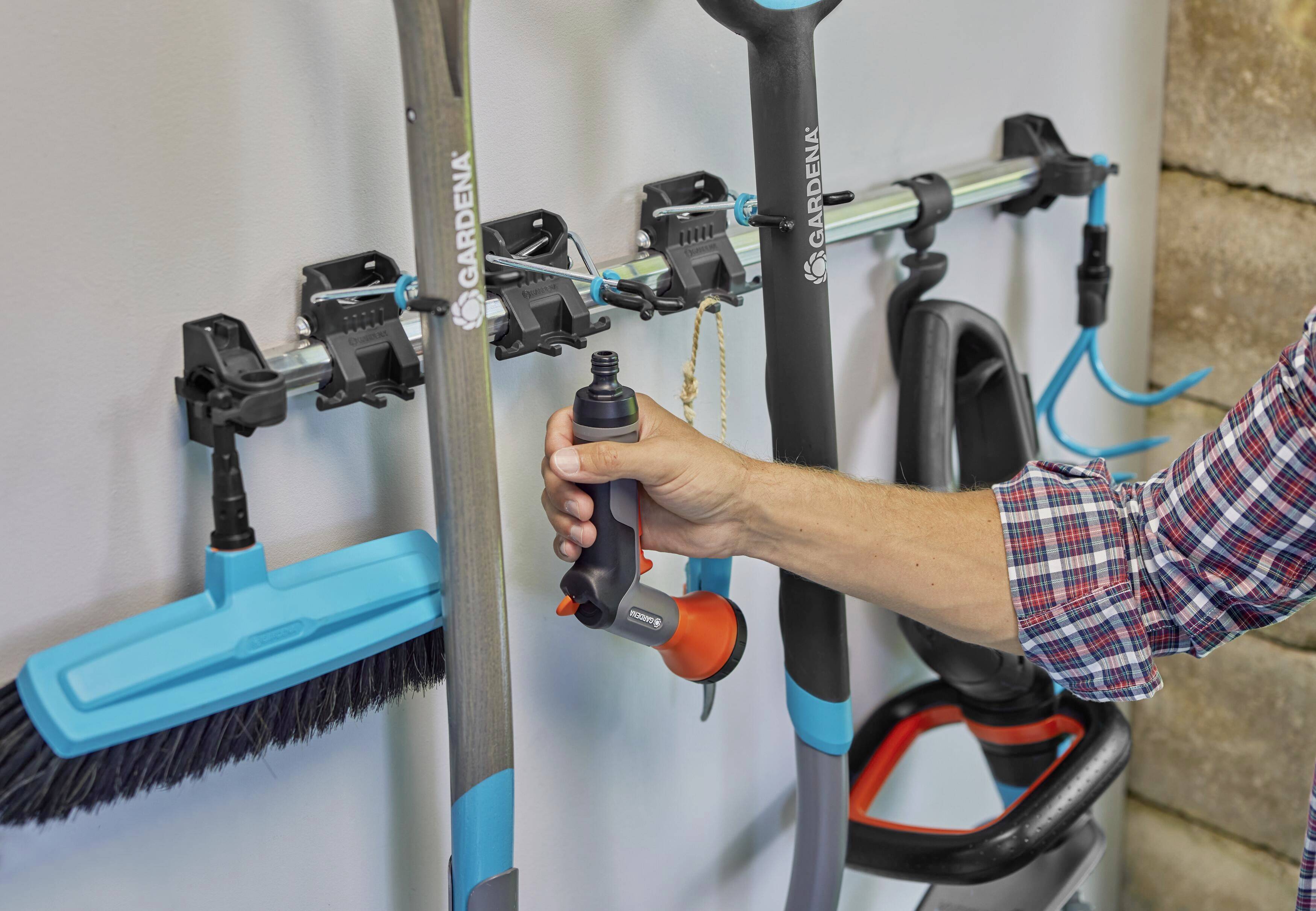 A wall-mounting system for garden tools shows a hand adjusting a handle on an orange device.