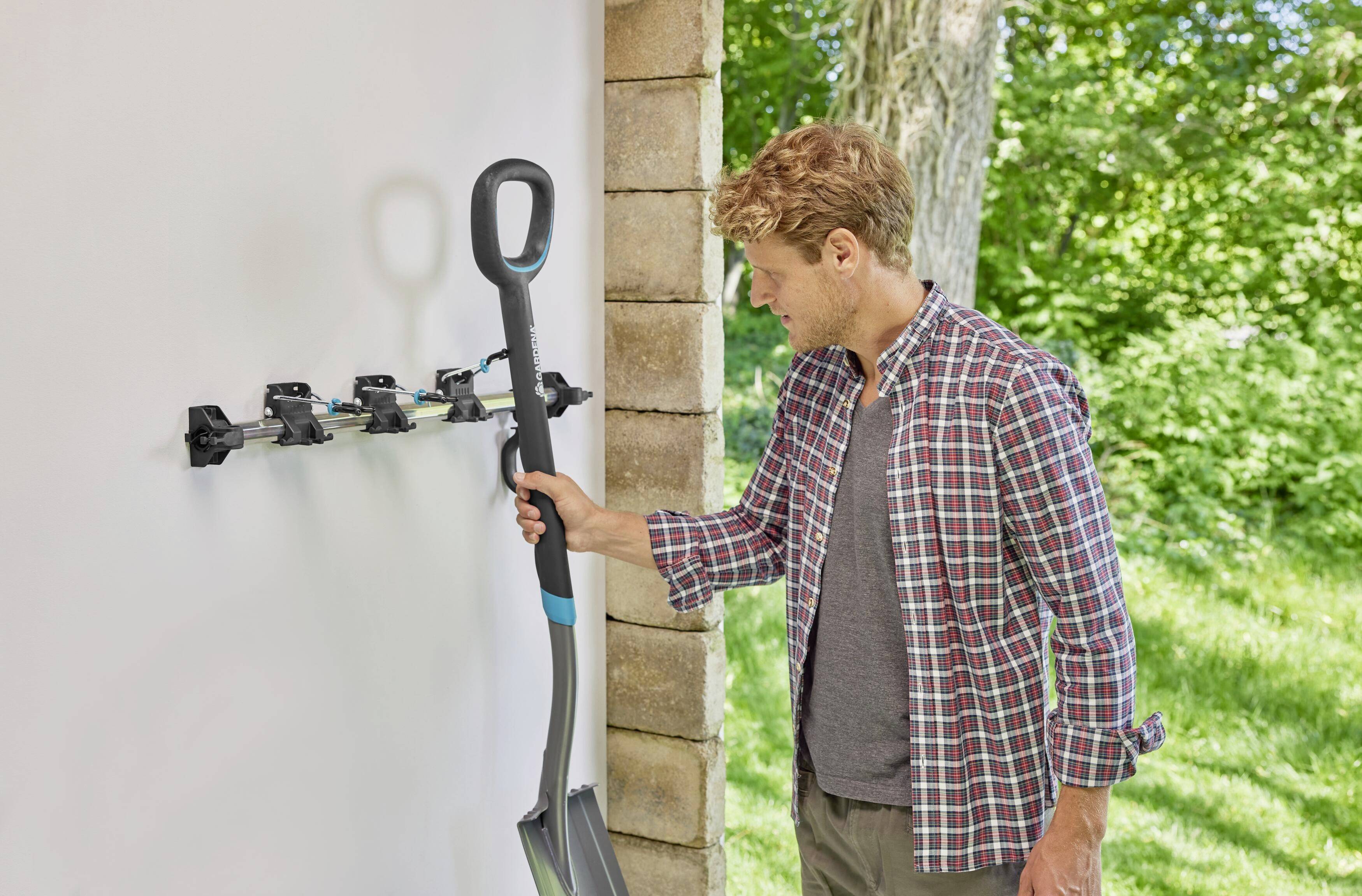 A man is attaching a spade to an outdoor wall mounting. Trees and a meadow are visible in the background.