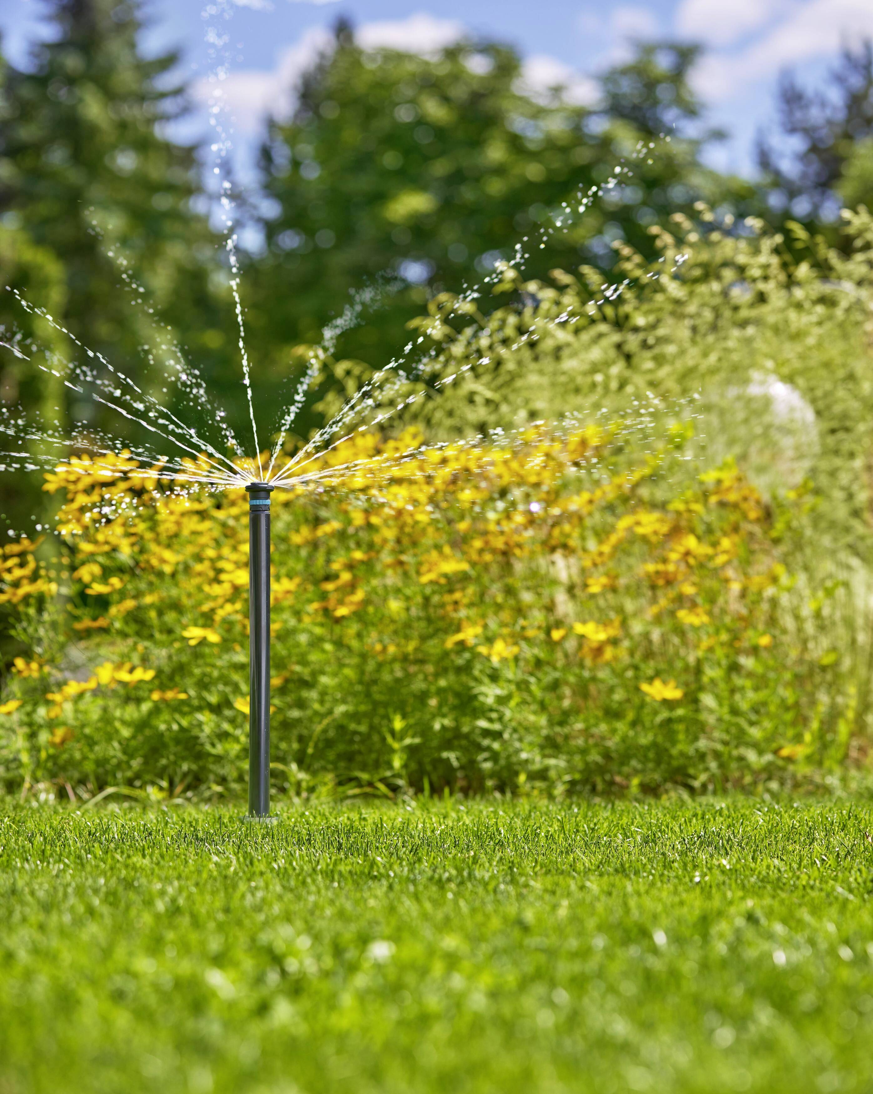 A lawn sprinkler with a fan-like design disperses water jets across a green lawn, with yellow flowers and trees in the background on a sunny day.