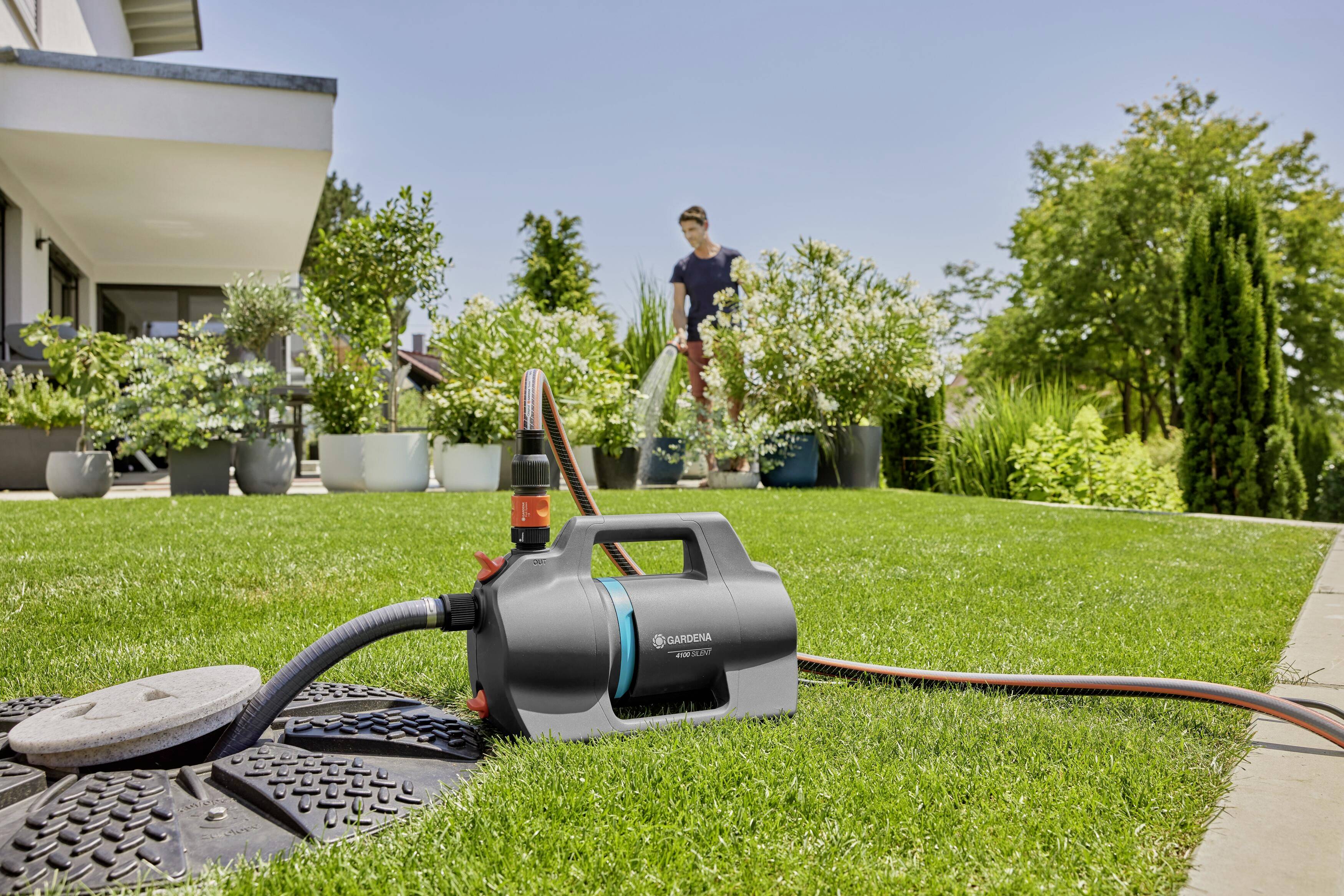 A water pump on a lawn, connected to a hosepipe. In the background, a person is watering plants in the garden.