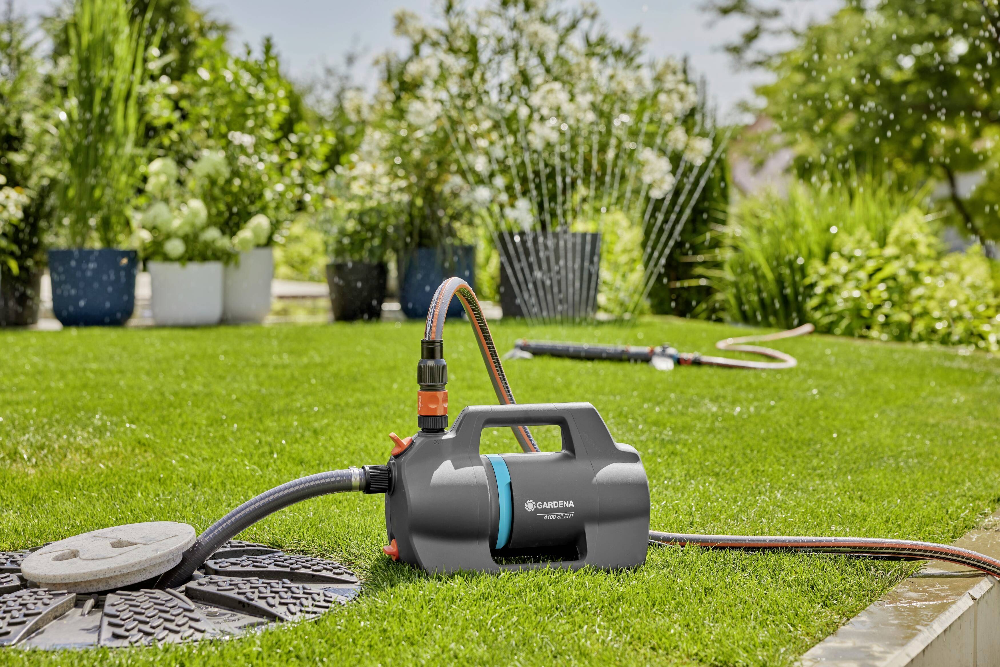 An electric garden pump is watering a lawn. In the background, several flowering plants are standing in pots, and a garden hose is visible.