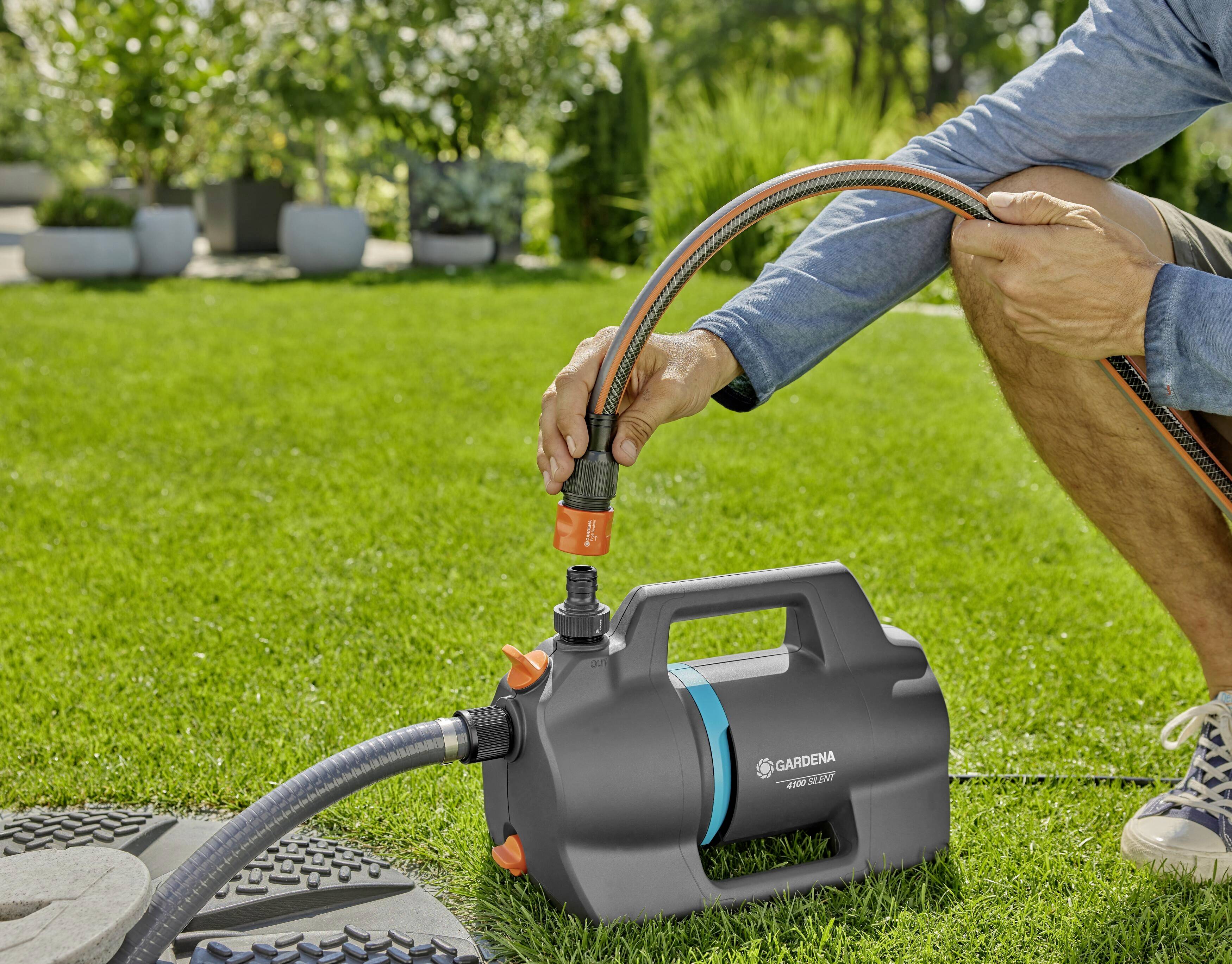 A person on grass is connecting a water hose to a black garden pump. Plants and trees are visible in the background.