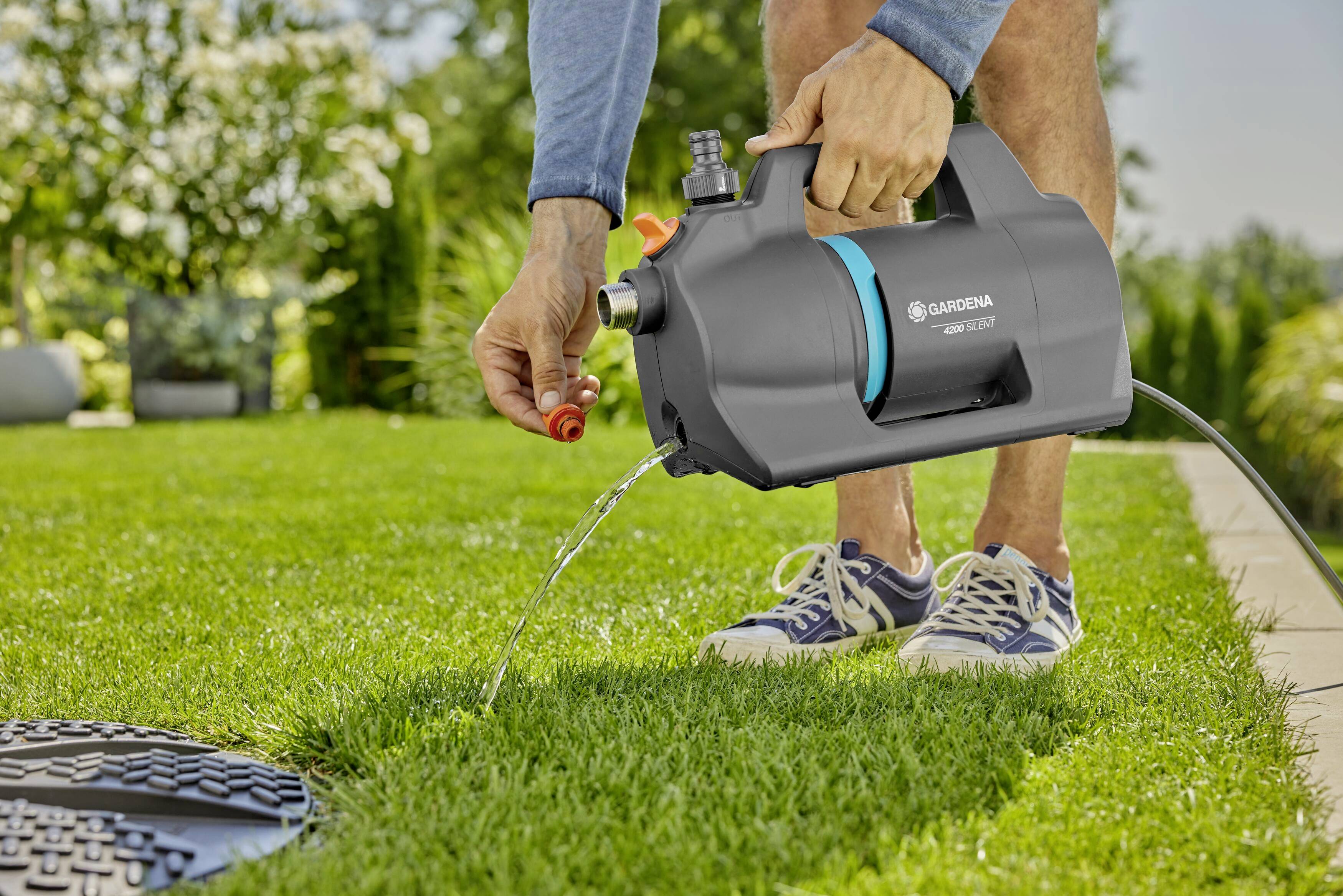 A person is watering a lawn with a portable garden sprayer in the garden. The sprayer has a grey housing and a distinctive blue band.