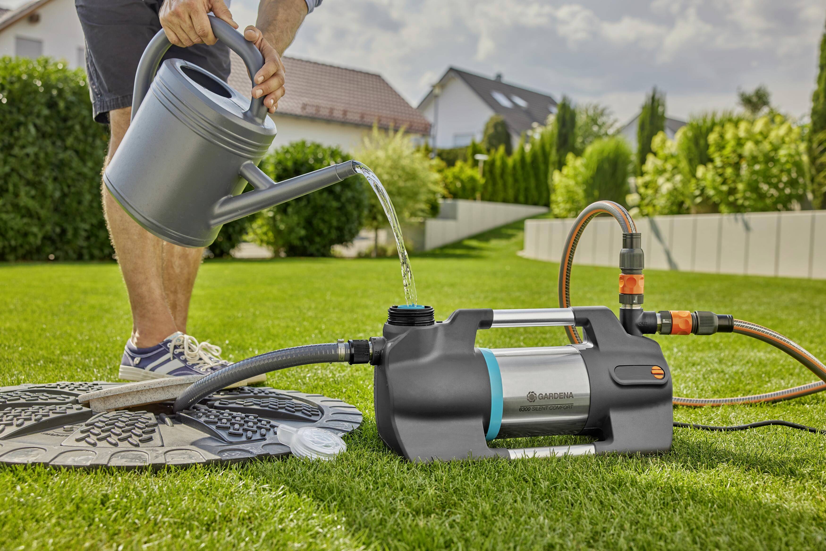 A person is pouring water into a watering can next to a water pump on a green lawn. The background shows houses and trees.