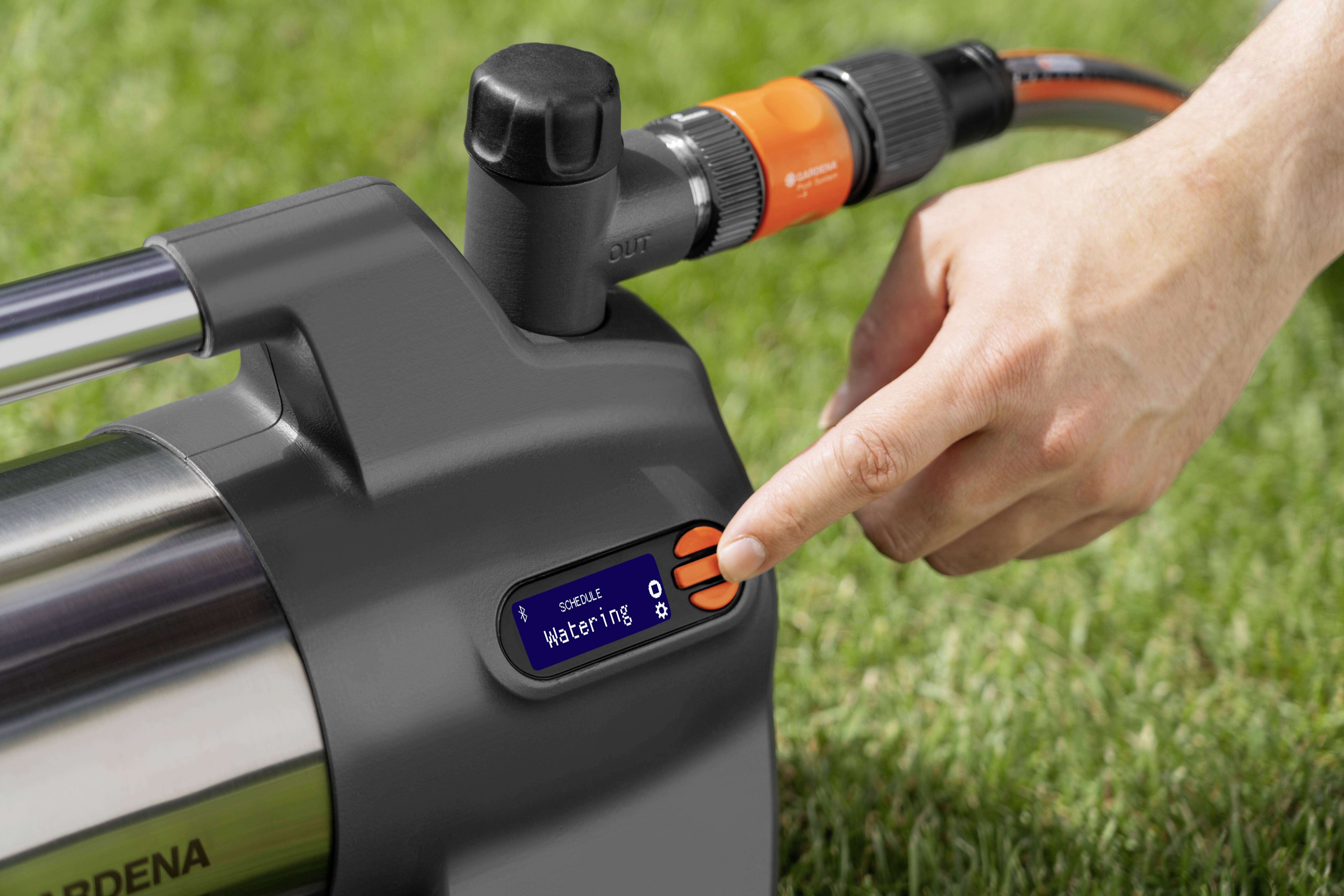 A finger presses a button on an irrigation computer situated on a lawn. The display shows 'Watering'.