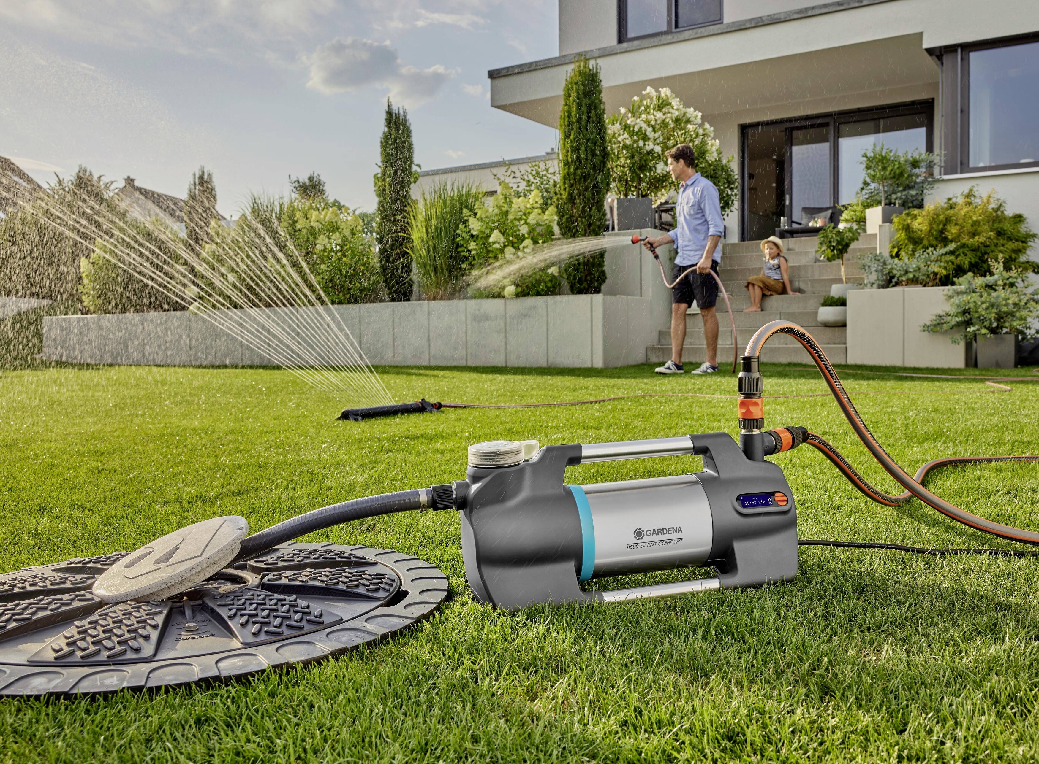 A man and a child are watering a lawn in the garden with a sprinkler and a garden hose. An irrigation system is visible.