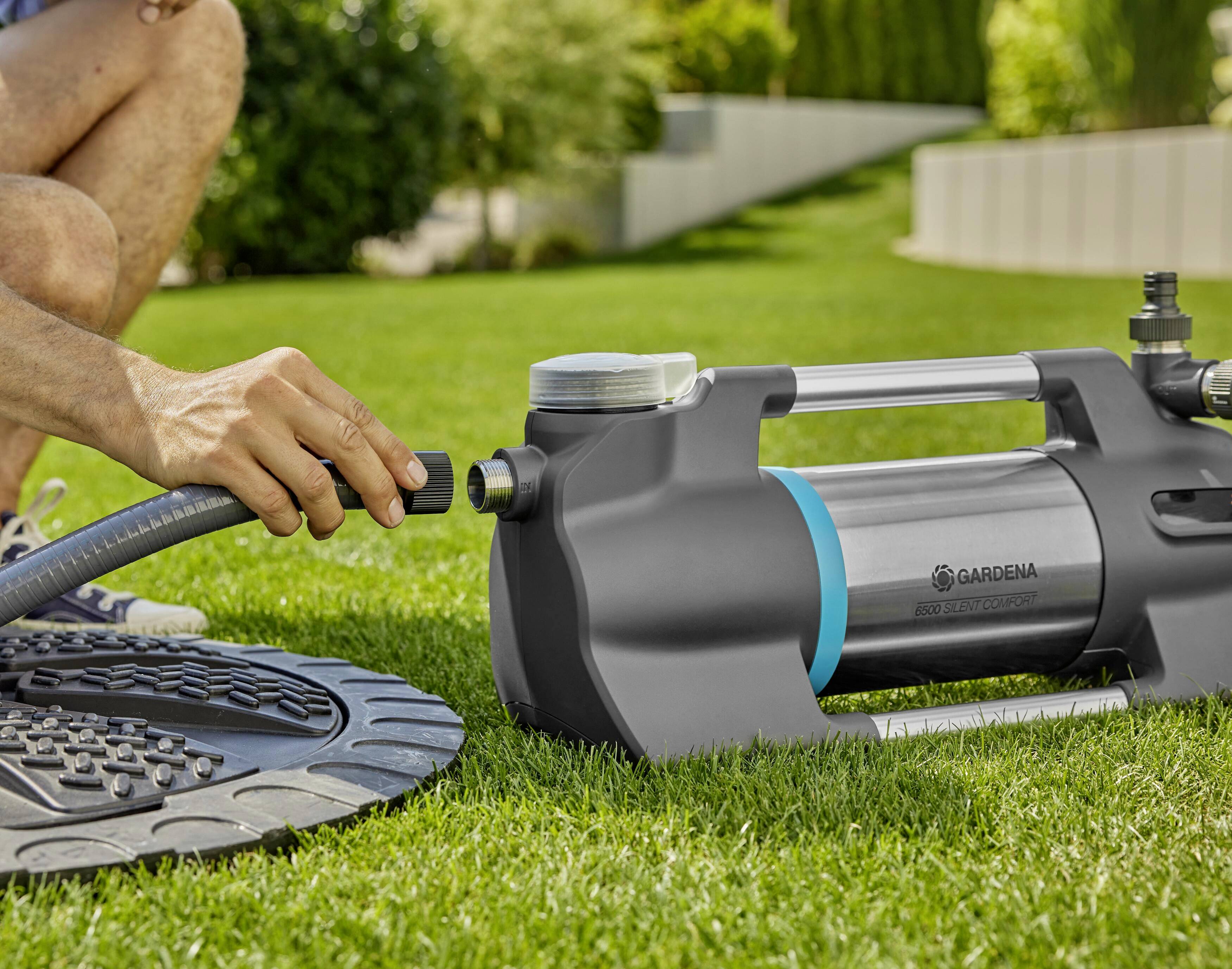 A person is connecting a hose to a garden pump on a lawn. Further garden areas can be seen in the background.