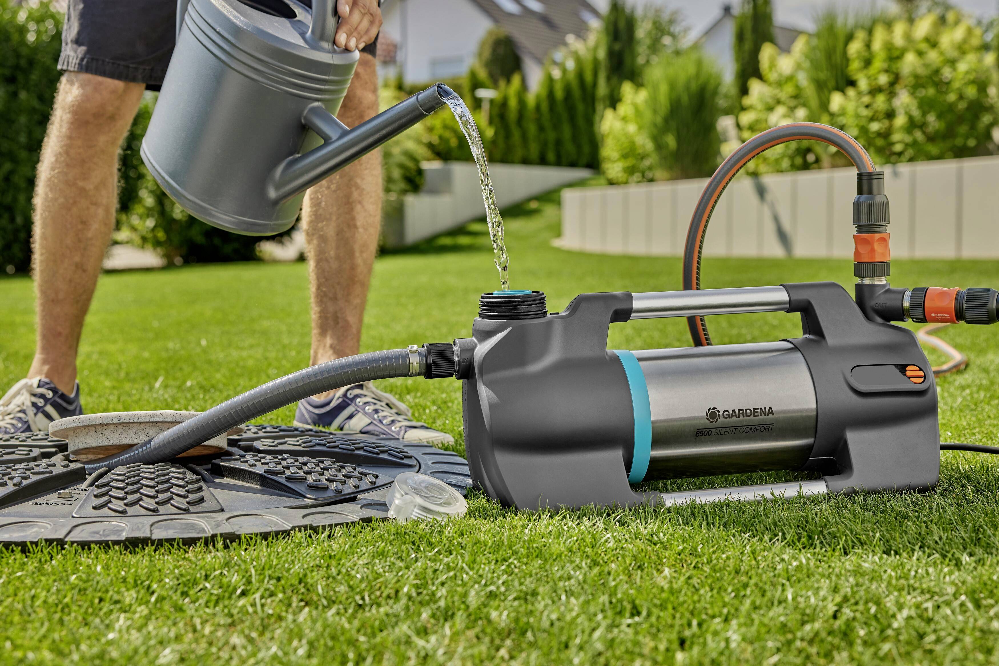 A person is pouring water into a portable garden sprayer on a lawn. The sprayer is connected to a hose and is standing next to a garden water tap.