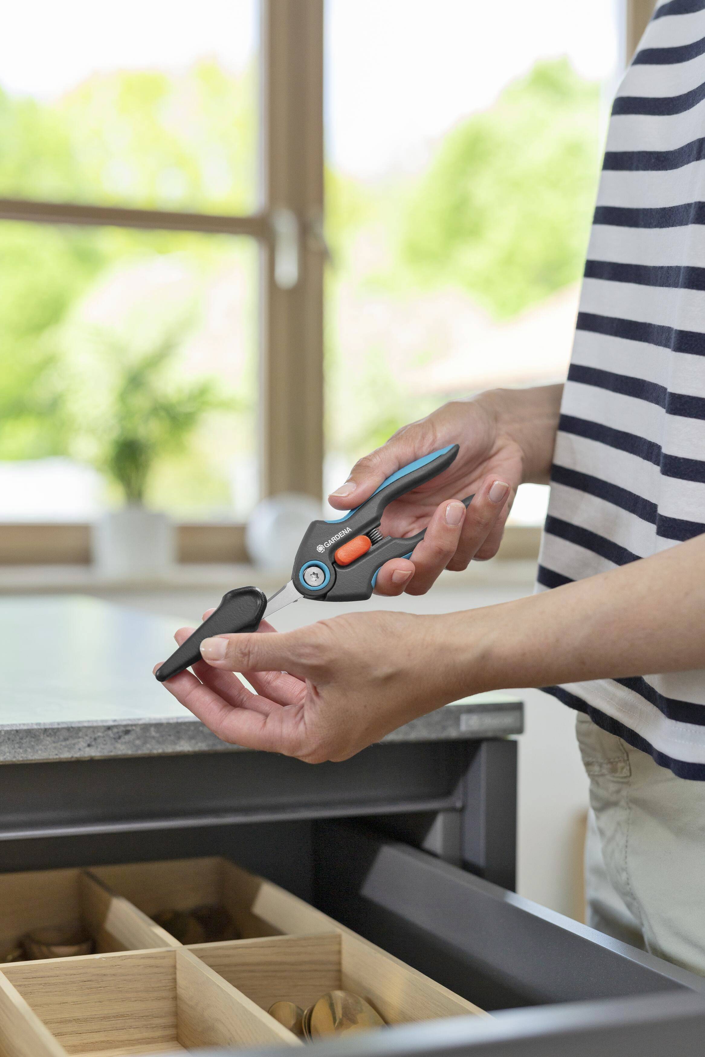 A person is holding kitchen scissors over an open drawer in a bright kitchen. Windows and plants are visible in the background.