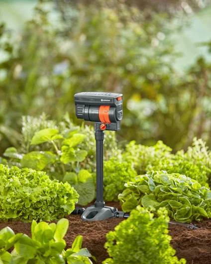 A compact sprinkler with a black and orange design is positioned among lush green plants in a garden, providing irrigation.
