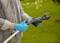 A person is pruning with secateurs in the garden. Gloves protect their hands. Green spaces and trees are visible in the background.