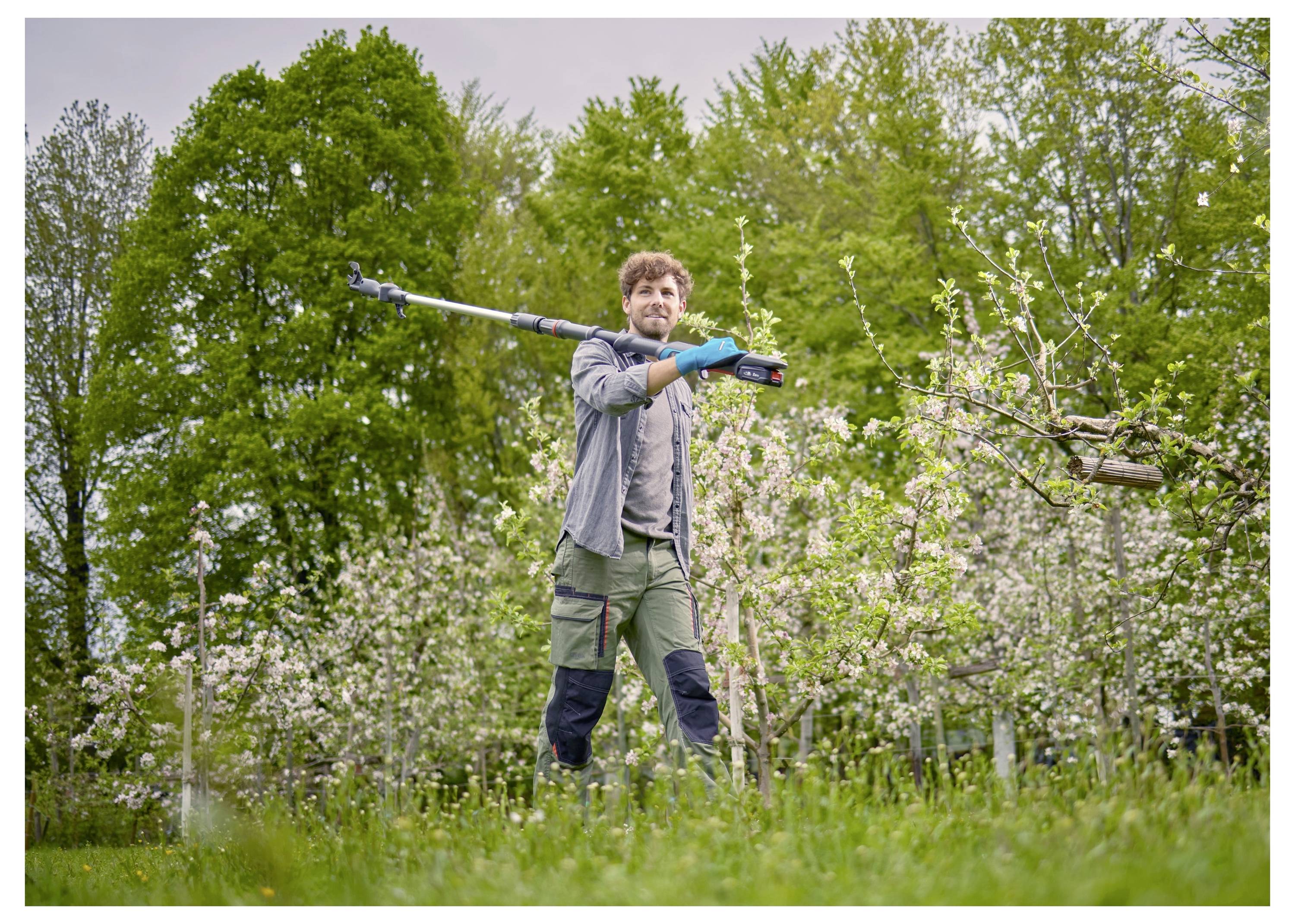 A person smiles while walking through an orchard, holding a pruning tool. Blossoming trees and lush greenery surround the scene.