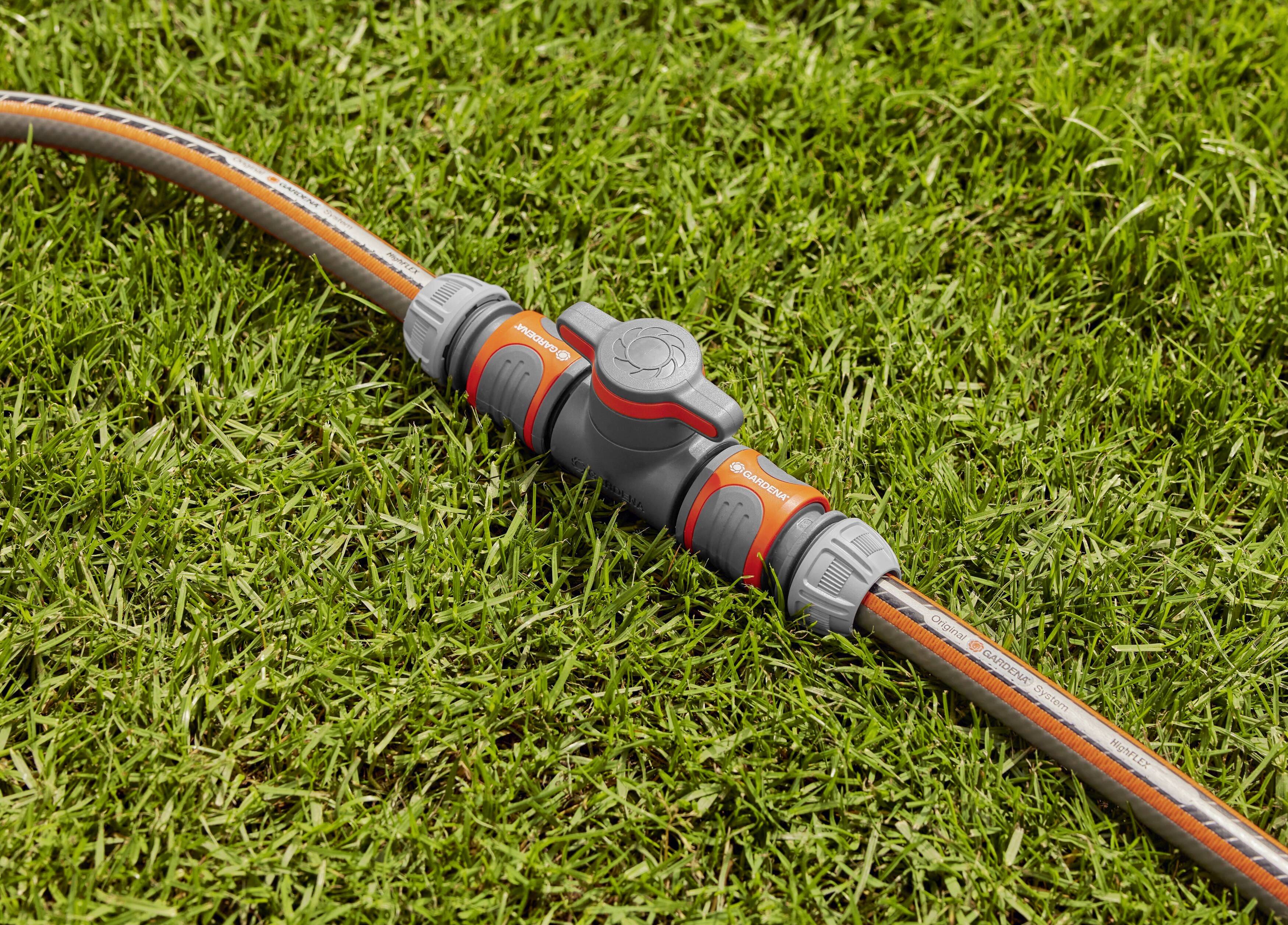A grey garden hose connector with a red lever is lying on green grass. The connector joins two hoses together.