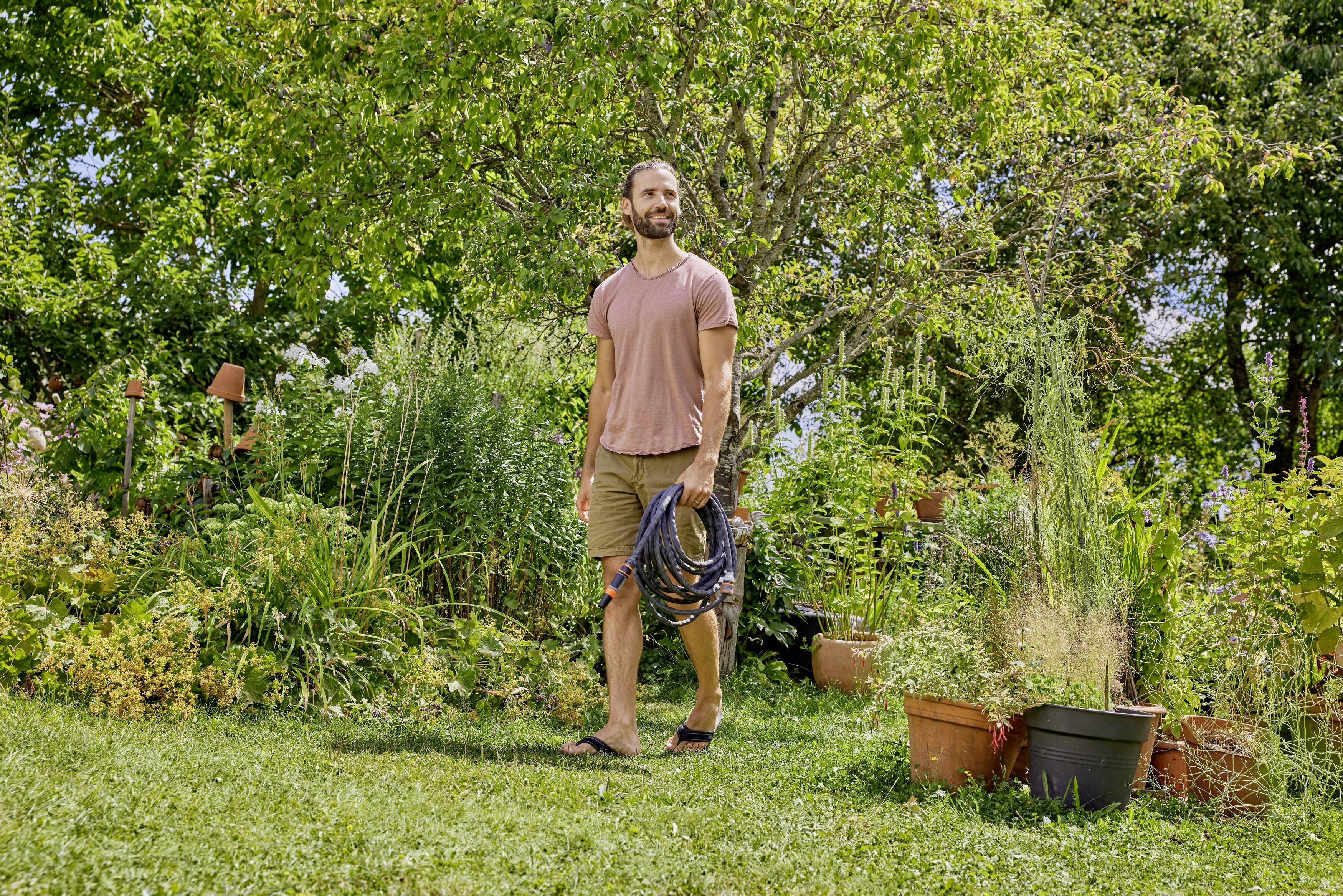 A man with a hosepipe walks through a lush garden filled with numerous plants and trees, under sunny weather, with a relaxed atmosphere.