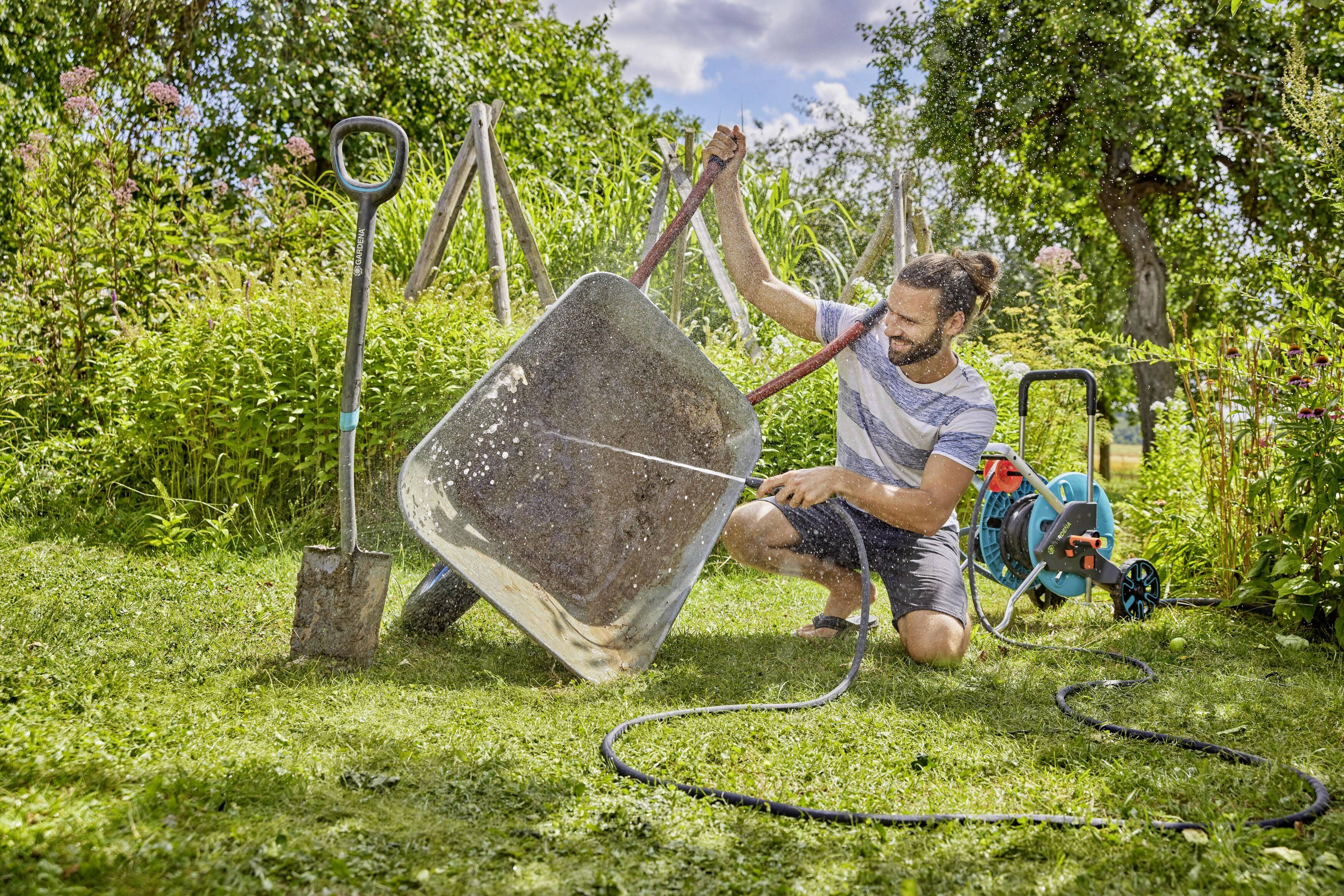 A person is cleaning a wheelbarrow with a hosepipe in the garden. A shovel is standing next to the wheelbarrow. It is sunny.