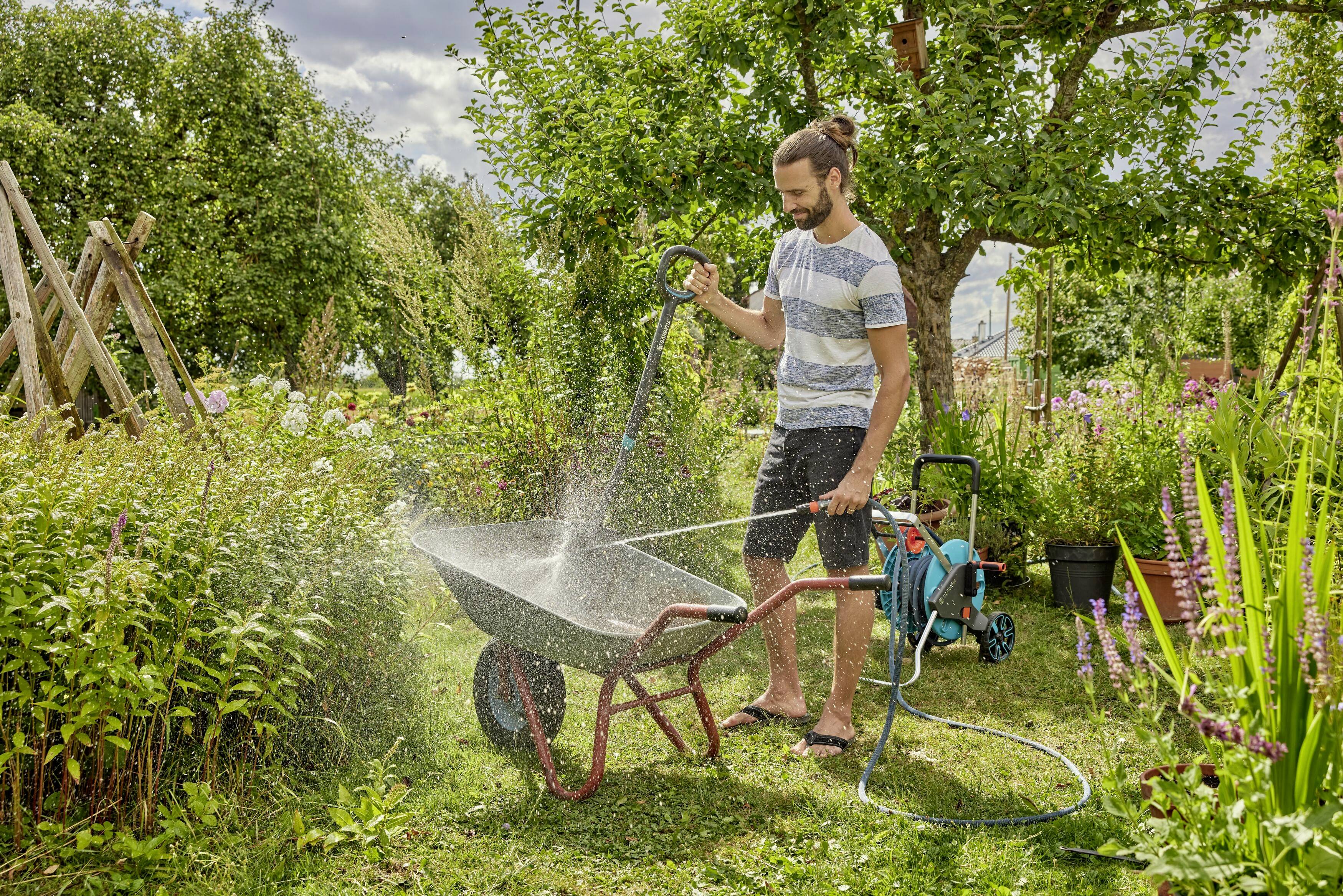A man is watering a wheelbarrow in the garden with a hosepipe, surrounded by plants and trees on a sunny day.