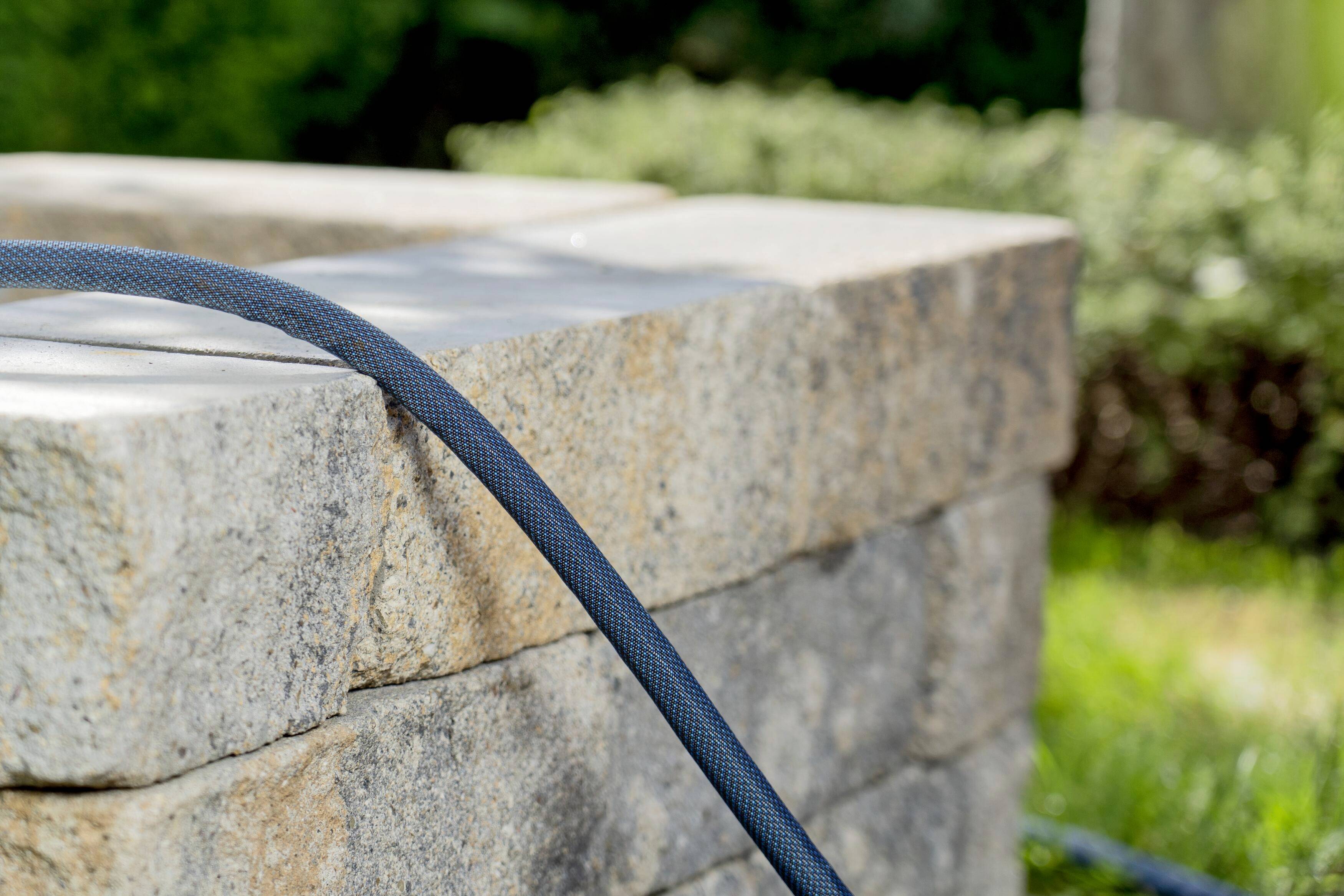 A grey garden hose lies on a low stone wall, surrounded by green grass and bushes in the background.