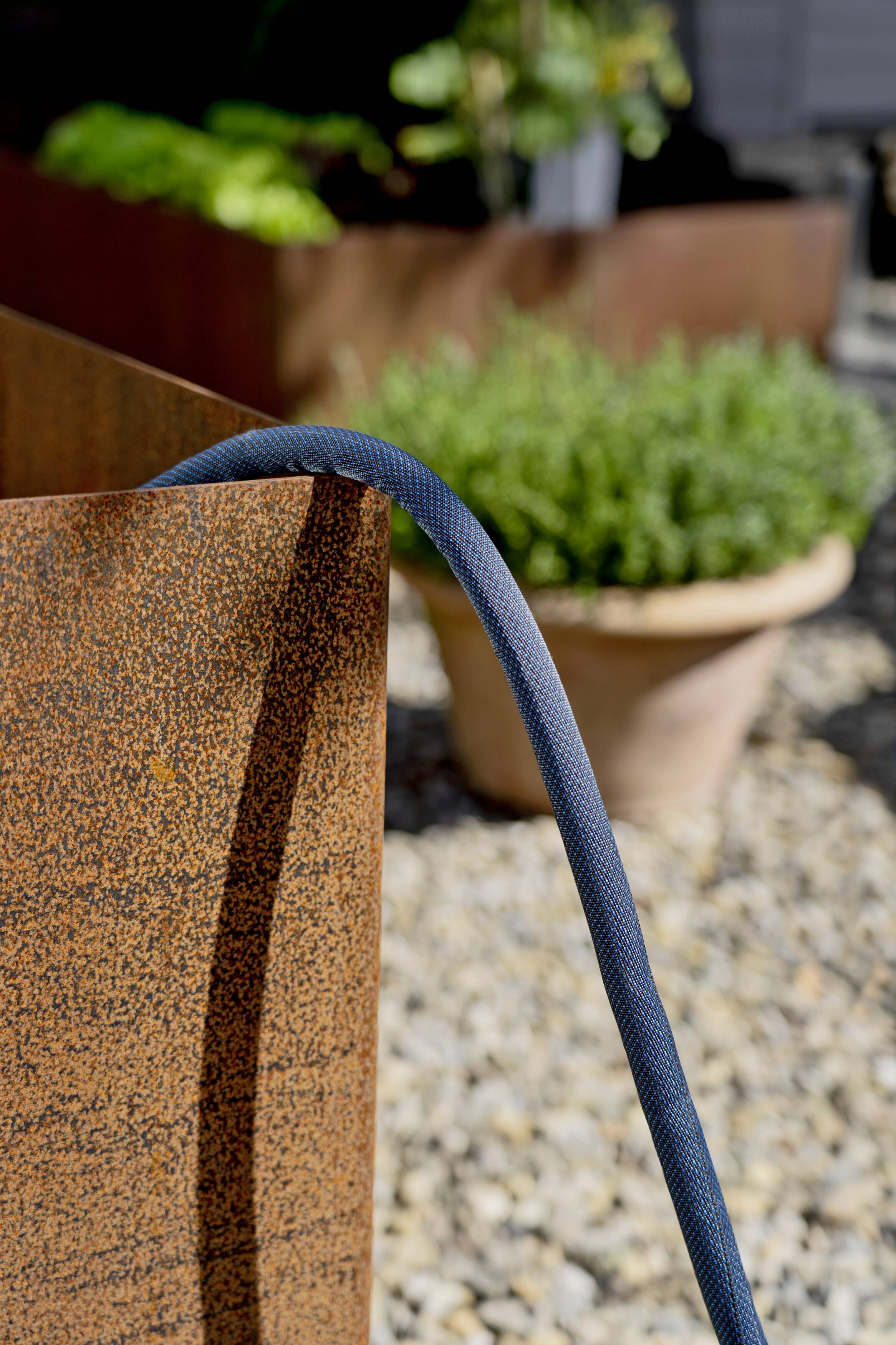 A blue garden hose hangs over a rusty metal container. In the background, a plant pot and pebbles are visible.