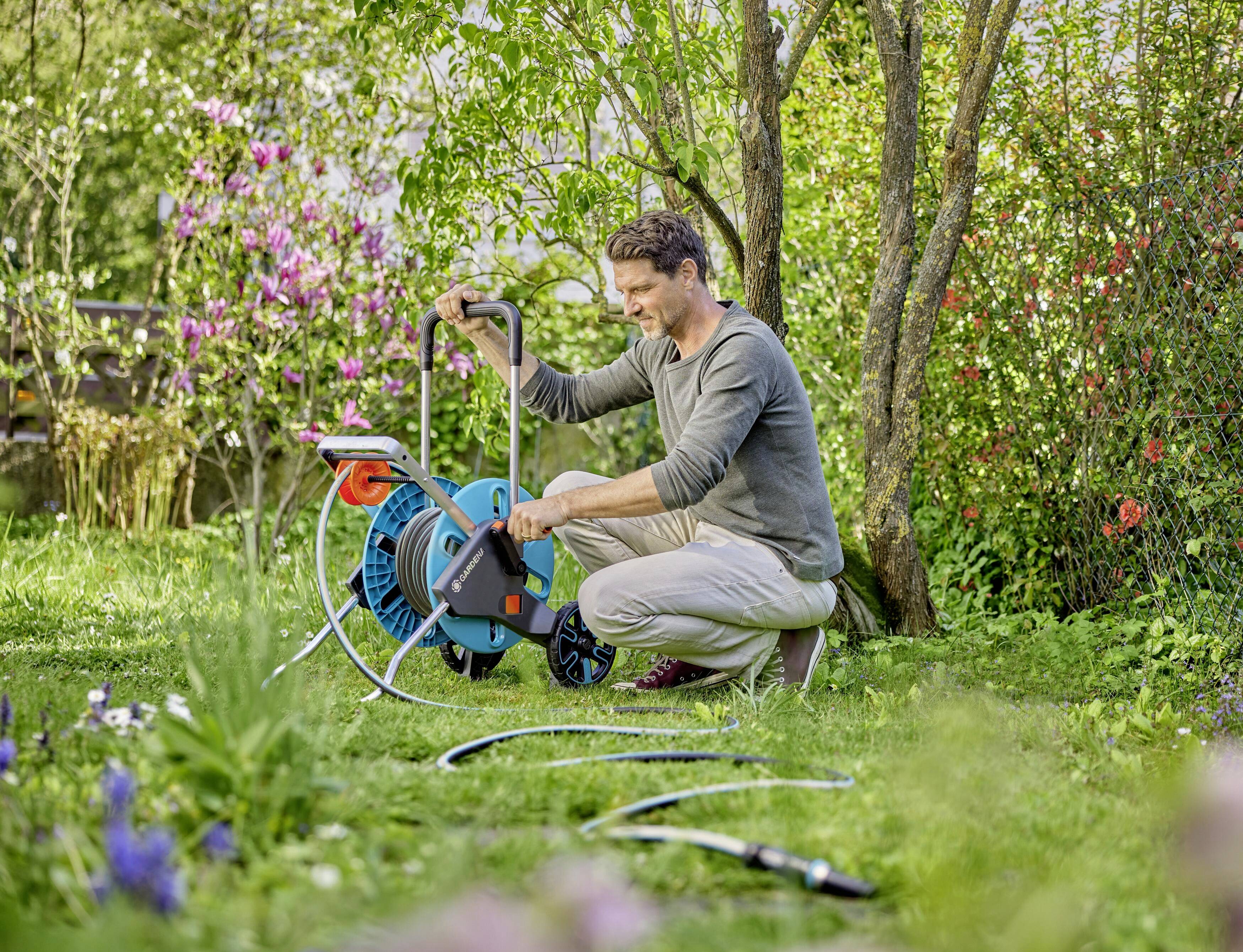 A person in the garden is unwinding the hose from a hose reel. Green plants and flowering bushes can be seen in the background.
