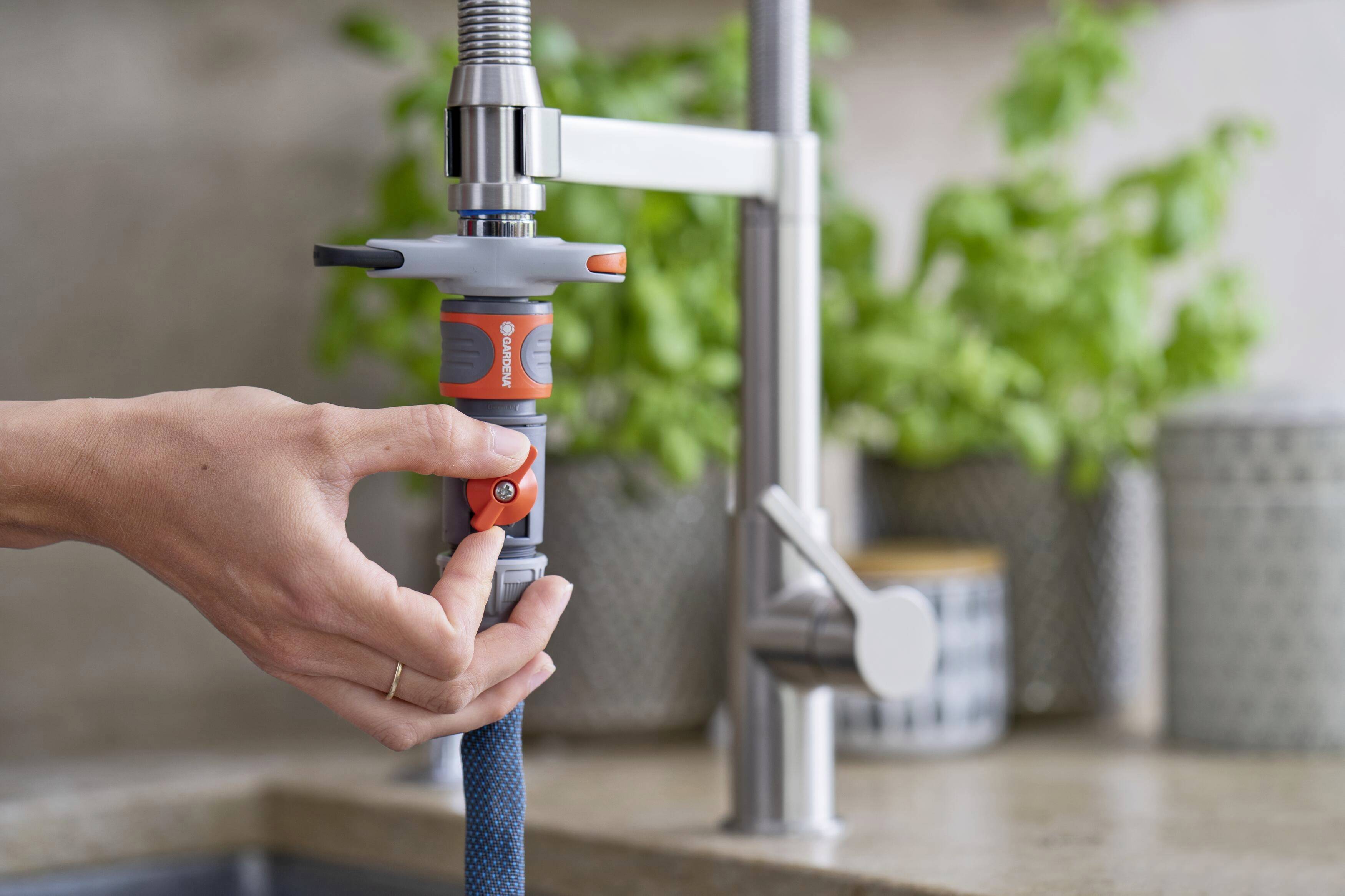 A hand is attaching a water hose to a kitchen tap adapter. Green plants can be seen on the worktop in the background.
