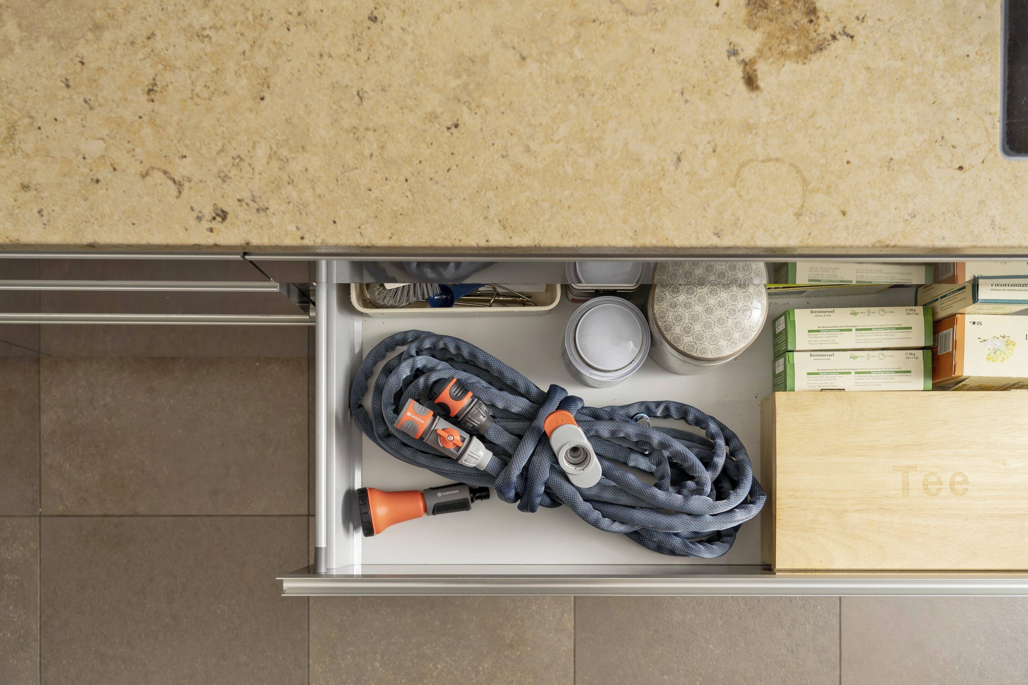 A kitchen drawer contains a garden hose with nozzles, two spice jars and a wooden box printed with 'Tea'.